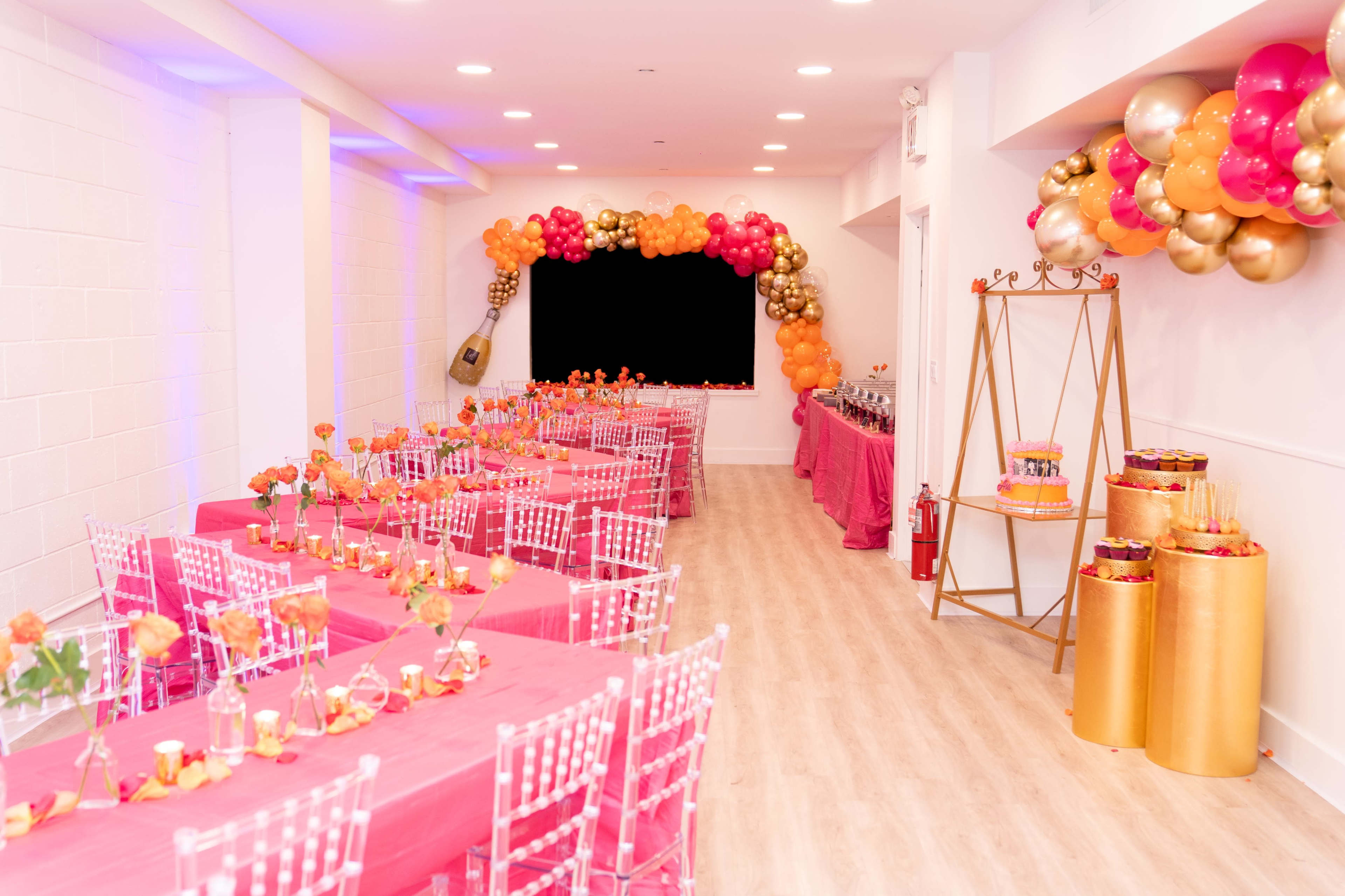 The image shows a decorated event space with pink tablecloths and floral centerpieces, featuring a snack table on the right side adorned with balloons and a golden display.