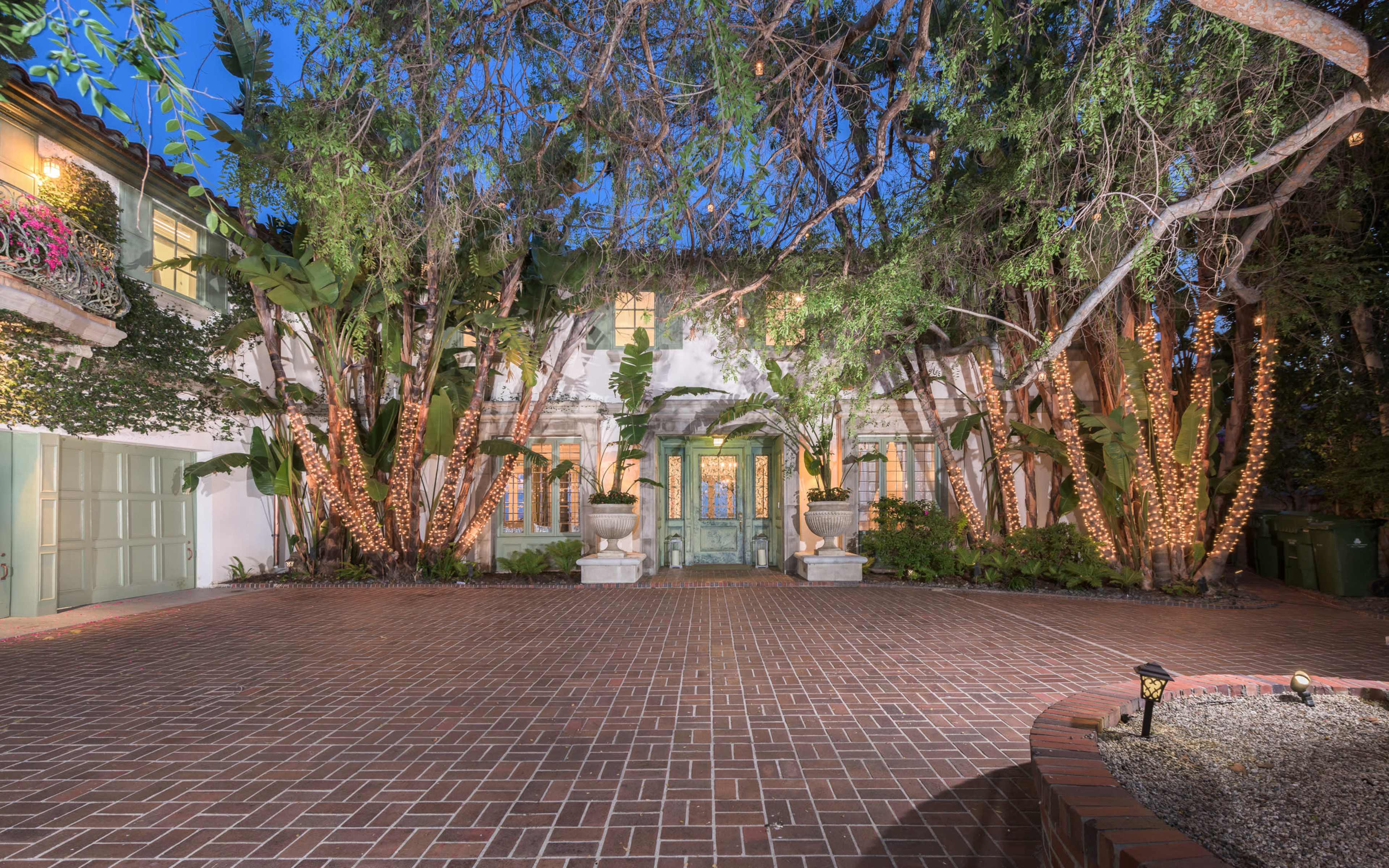 A large, two-story house with a green front door and surrounded by palm trees and illuminated by soft lights at dusk.