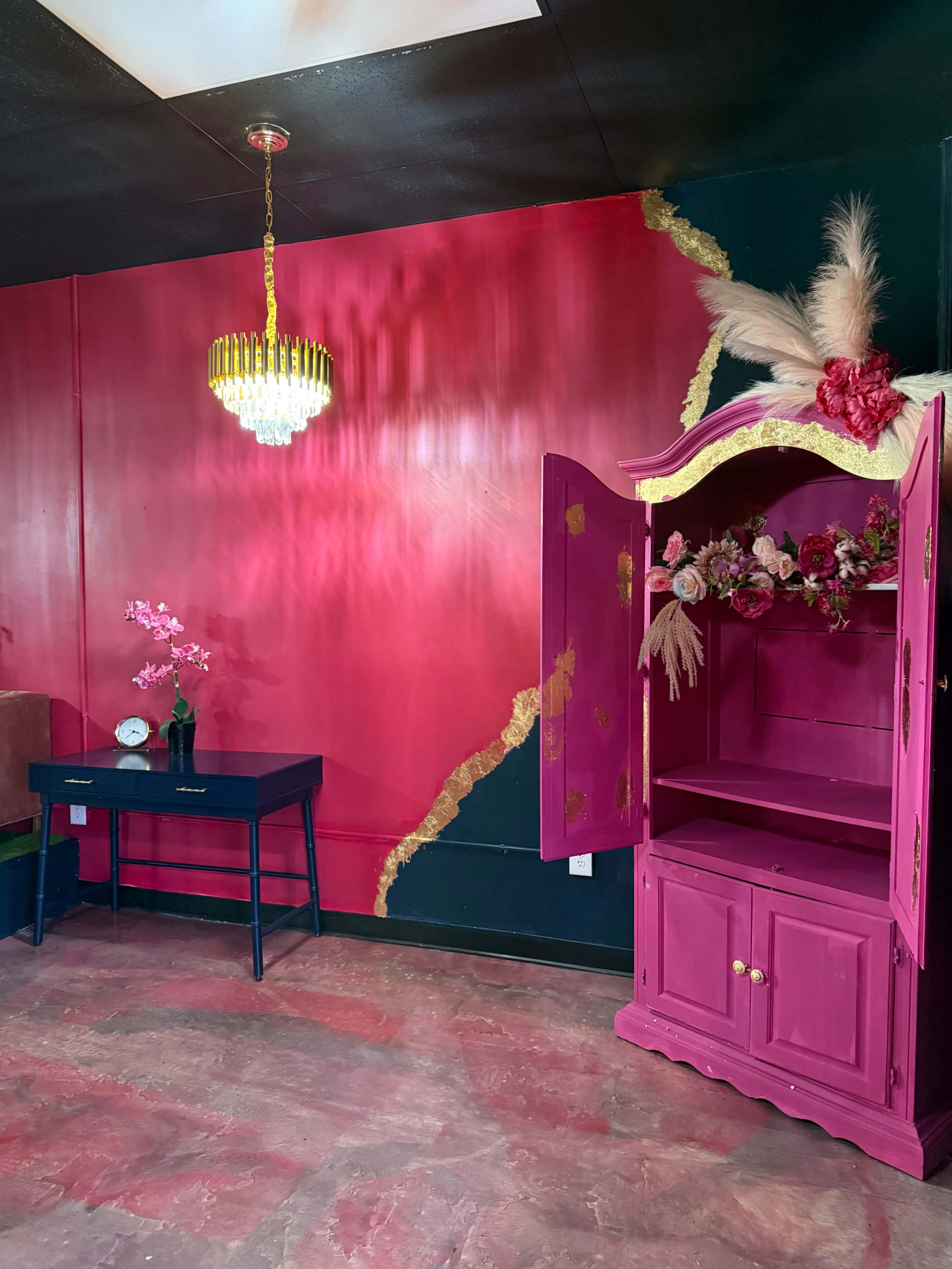 The scene features a vibrant pink wall with a gold accent, a crystal chandelier, a dark blue table with a clock, and a decorative pink cabinet adorned with flowers.