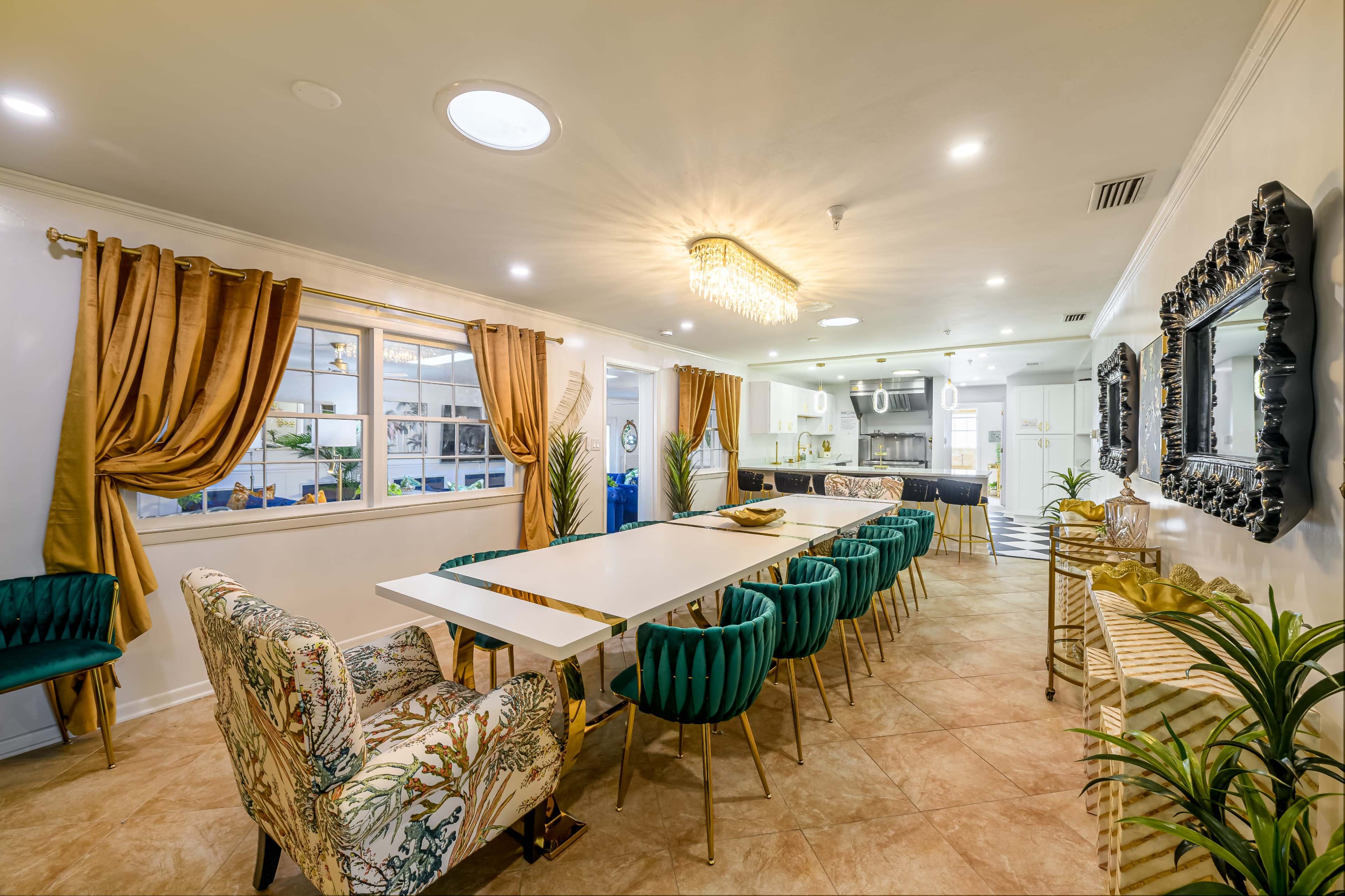 The image shows a dining area with a long white table surrounded by velvet green chairs, a floral patterned chair at one end, and large windows with golden drapes.