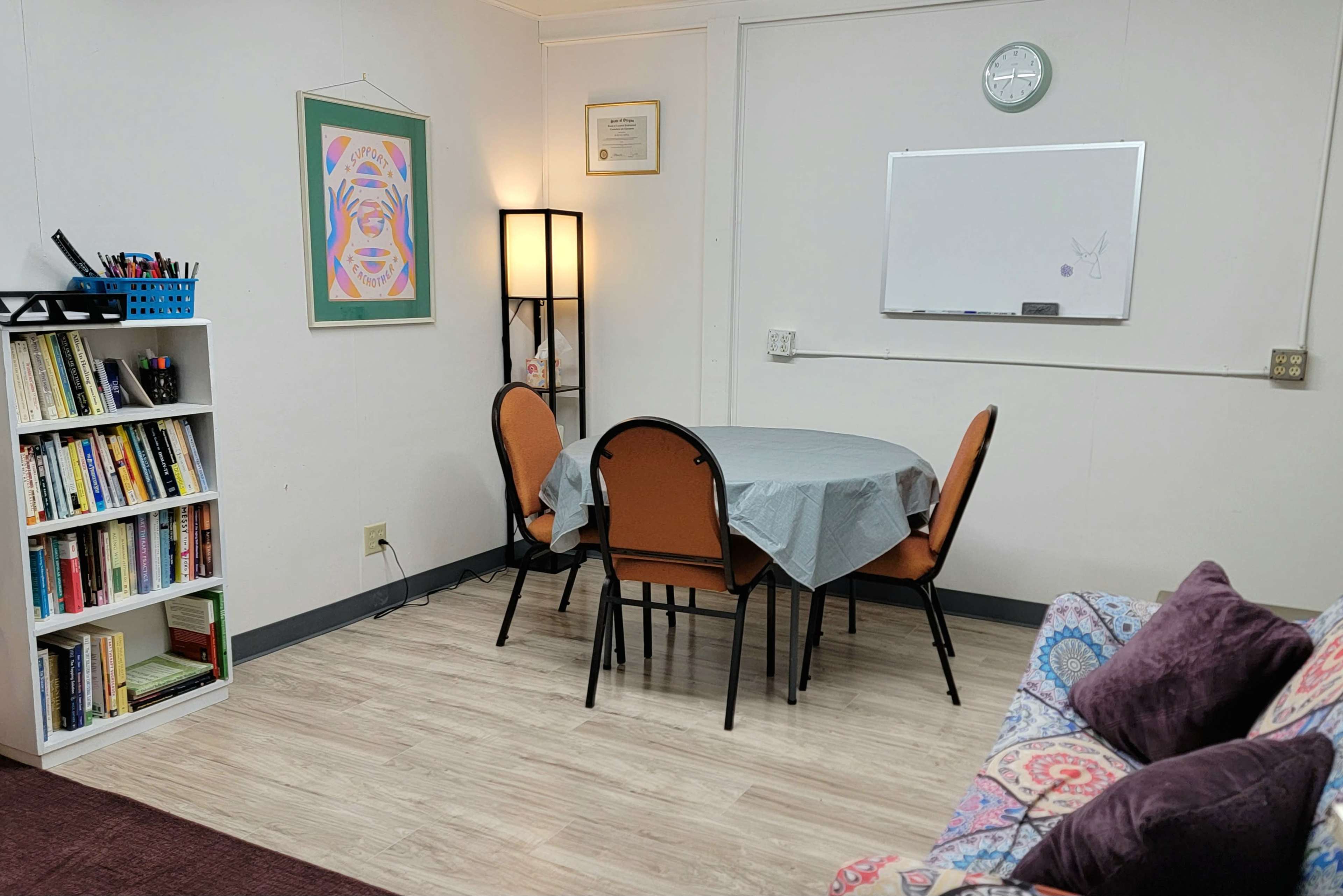 The image shows a simple room with a round table and four chairs covered by a gray tablecloth, a bookshelf filled with books, a framed artwork on the wall, a light fixture, and a whiteboard.