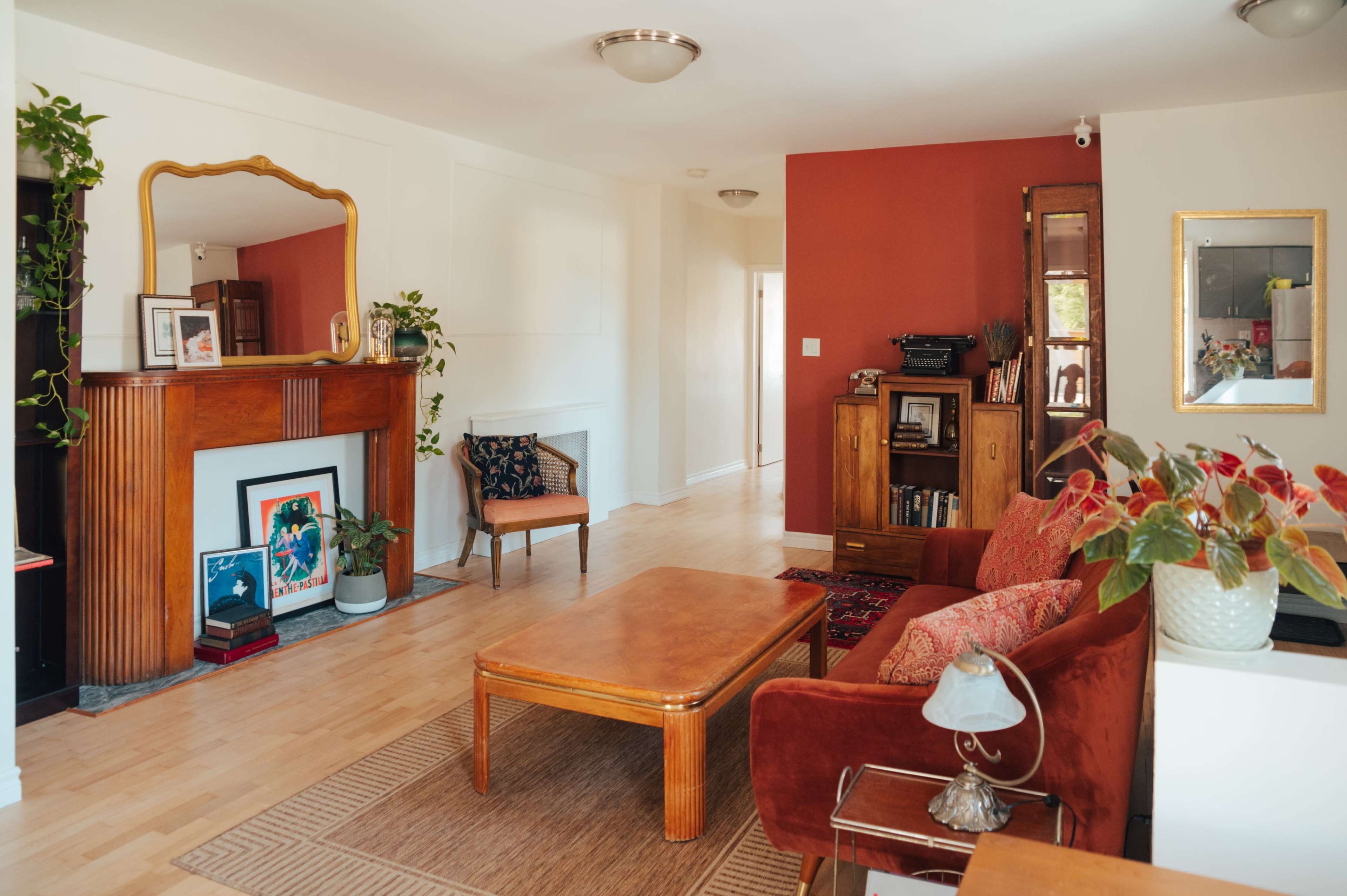 The image shows a living room featuring a red accent wall, a wooden fireplace with a mirror above it, a coffee table, and two red armchairs, complemented by plants and decor.
