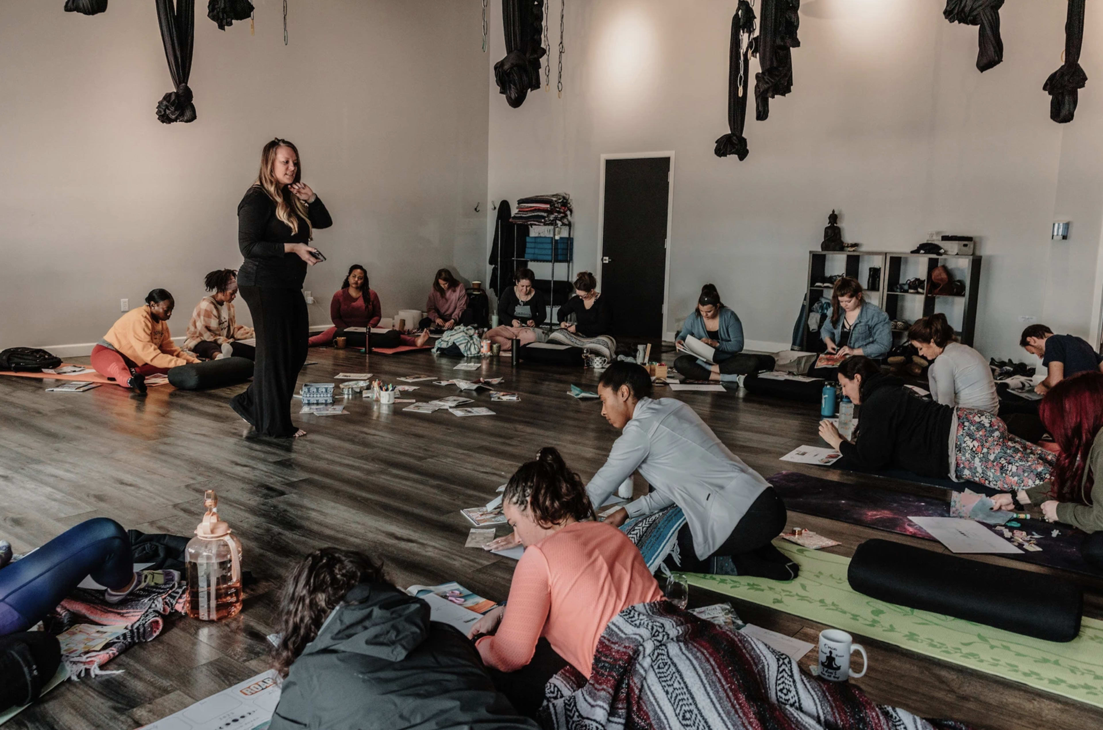A group of people sits on yoga mats in a studio, engaging in a workshop while a leader presents in the front.