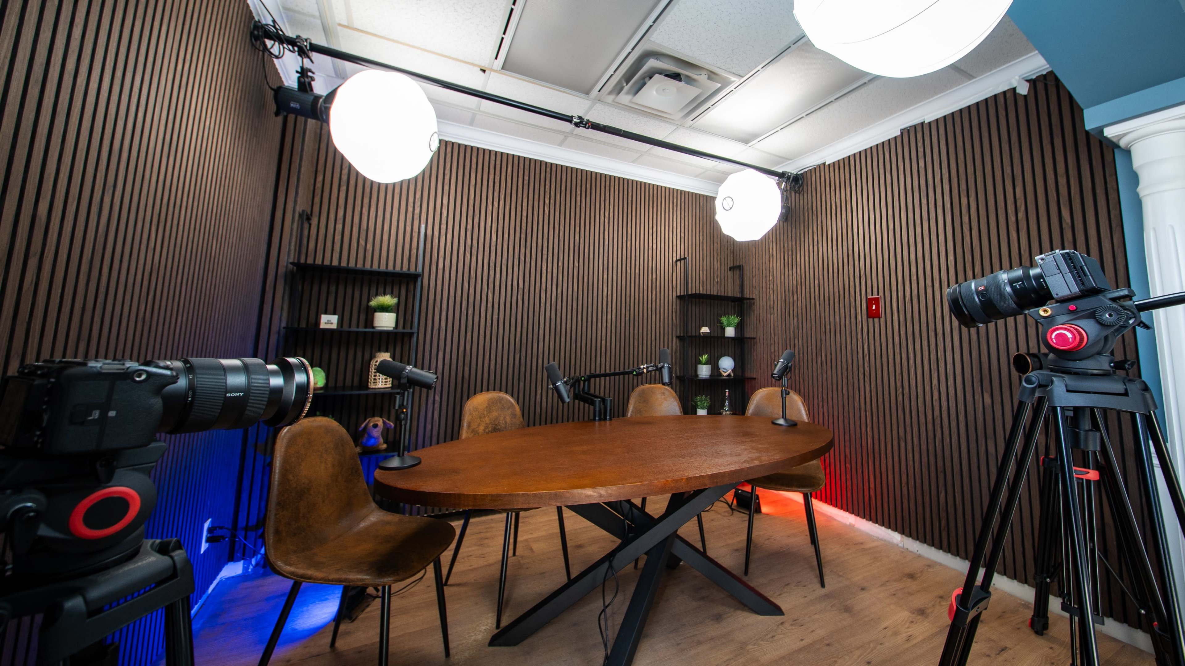 The image shows a podcast recording studio with a wooden table, four chairs, and three cameras set up on tripods, along with pendant lights and a wooden slatted wall.