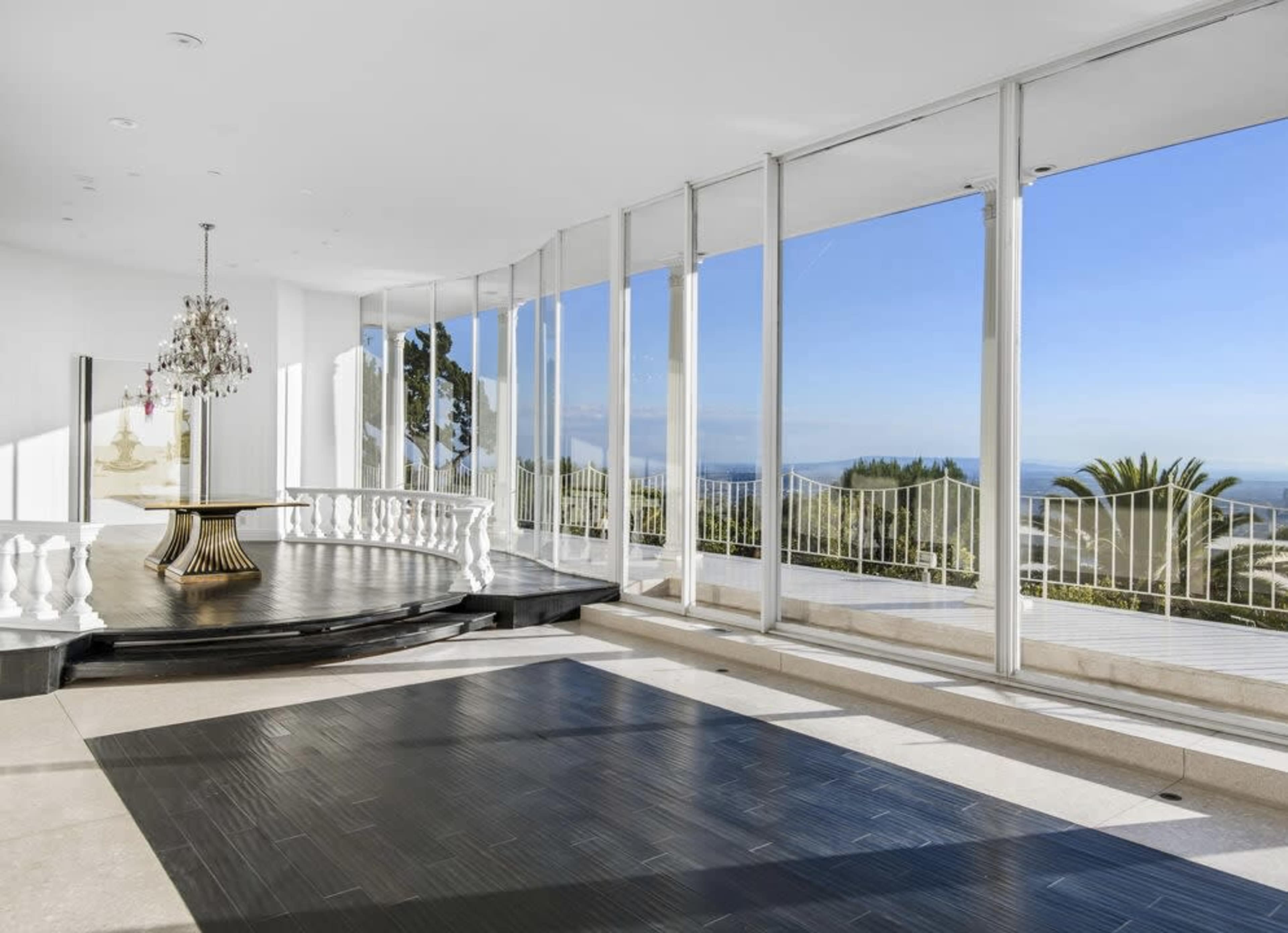The image shows an expansive room with large glass walls offering a view of the landscape, featuring a central circular dining table and a chandelier.