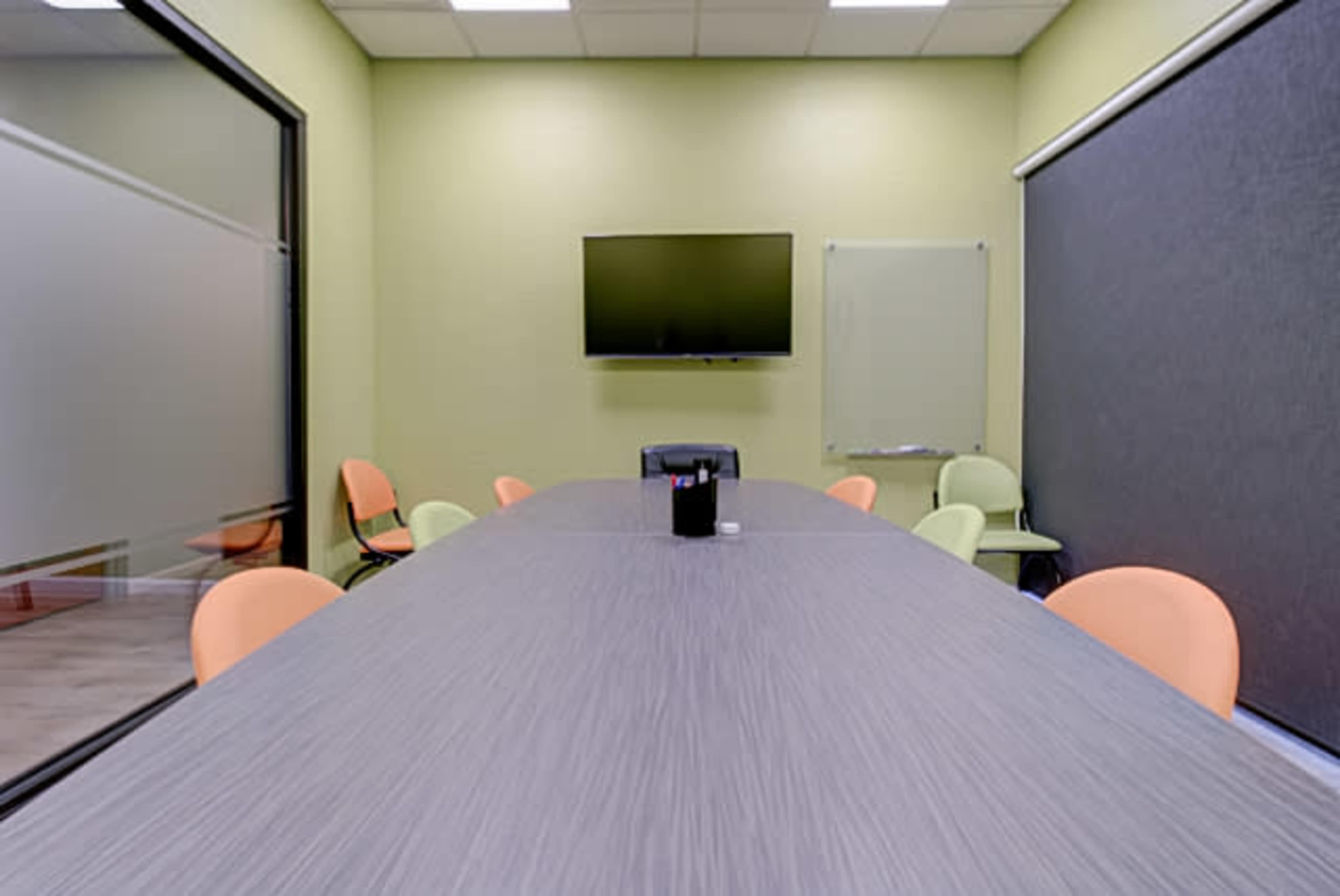 A conference room features a long gray table with orange chairs, a television mounted on the wall, and large glass panels.