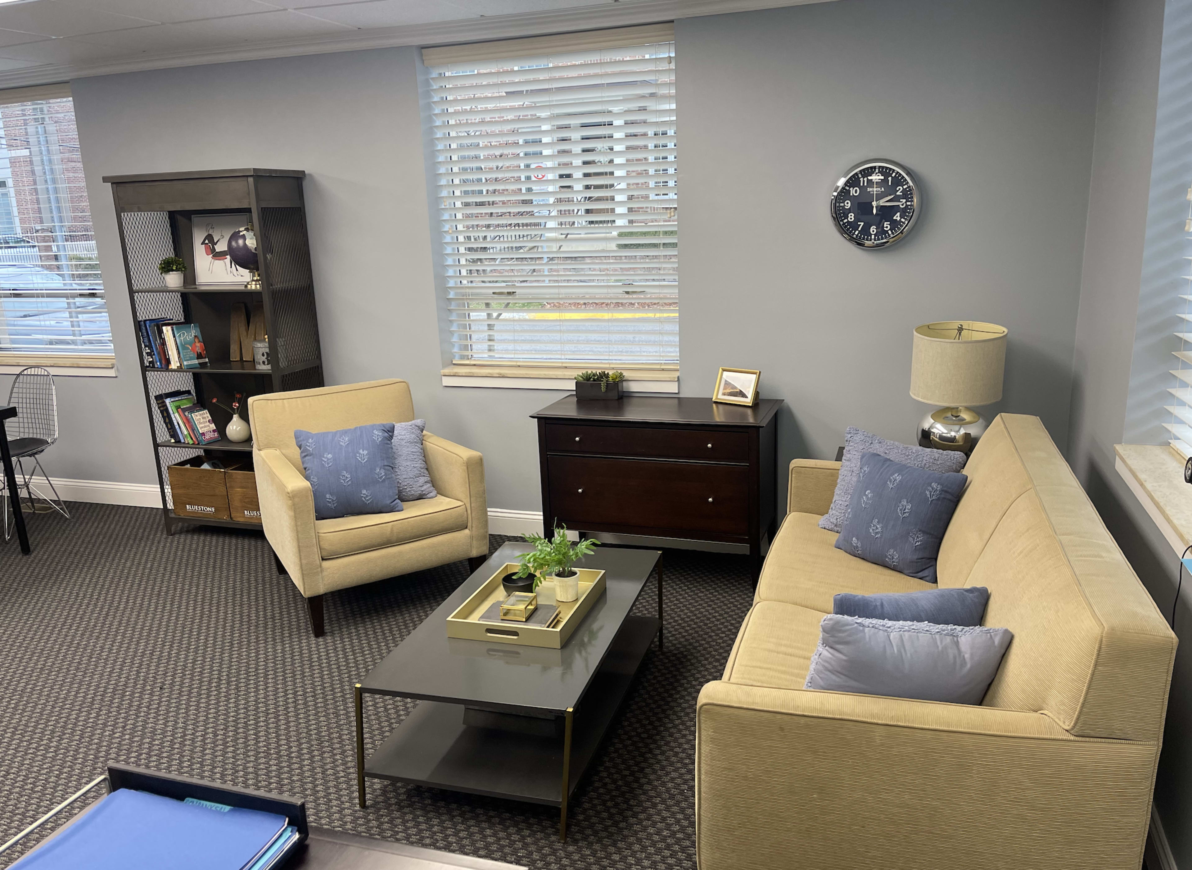 A modern office waiting area with a beige sofa and armchair, a black coffee table, a wooden bookshelf, and a wall clock.