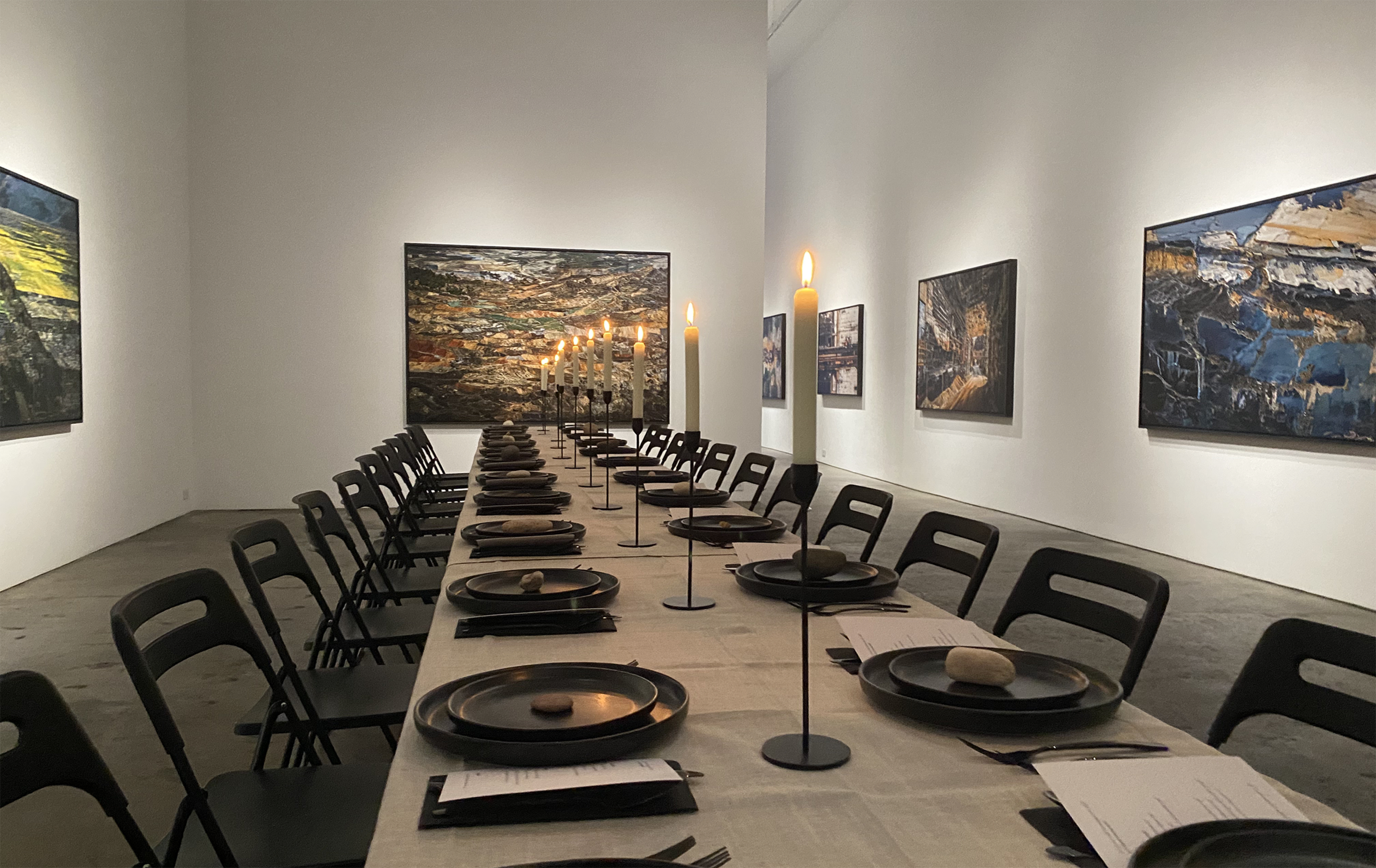A long, empty dining table is set in a gallery surrounded by various paintings on the walls.