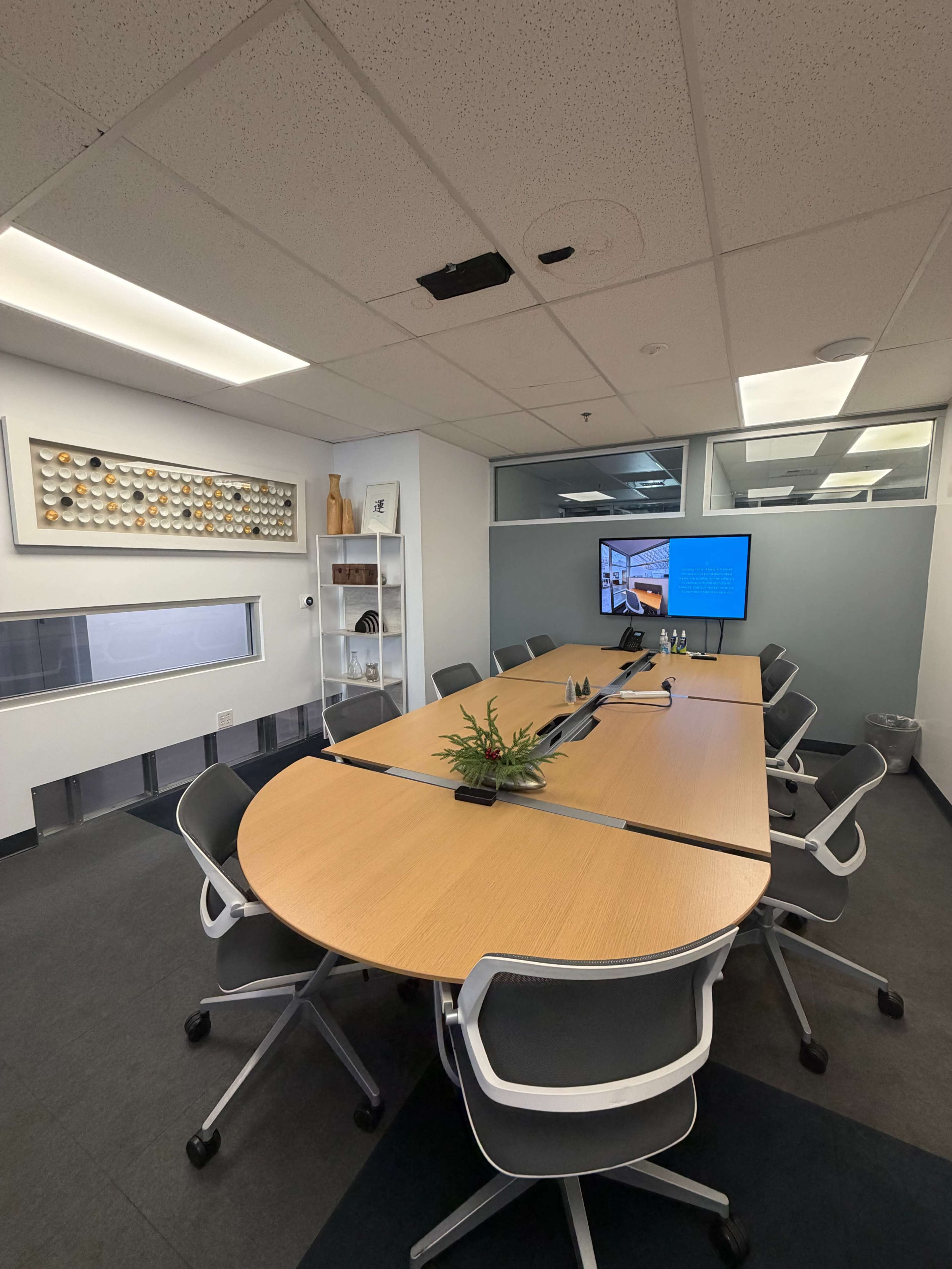 The image shows a modern conference room equipped with a large oval table, several chairs, a television screen displaying information, and a small shelf with decorative items.