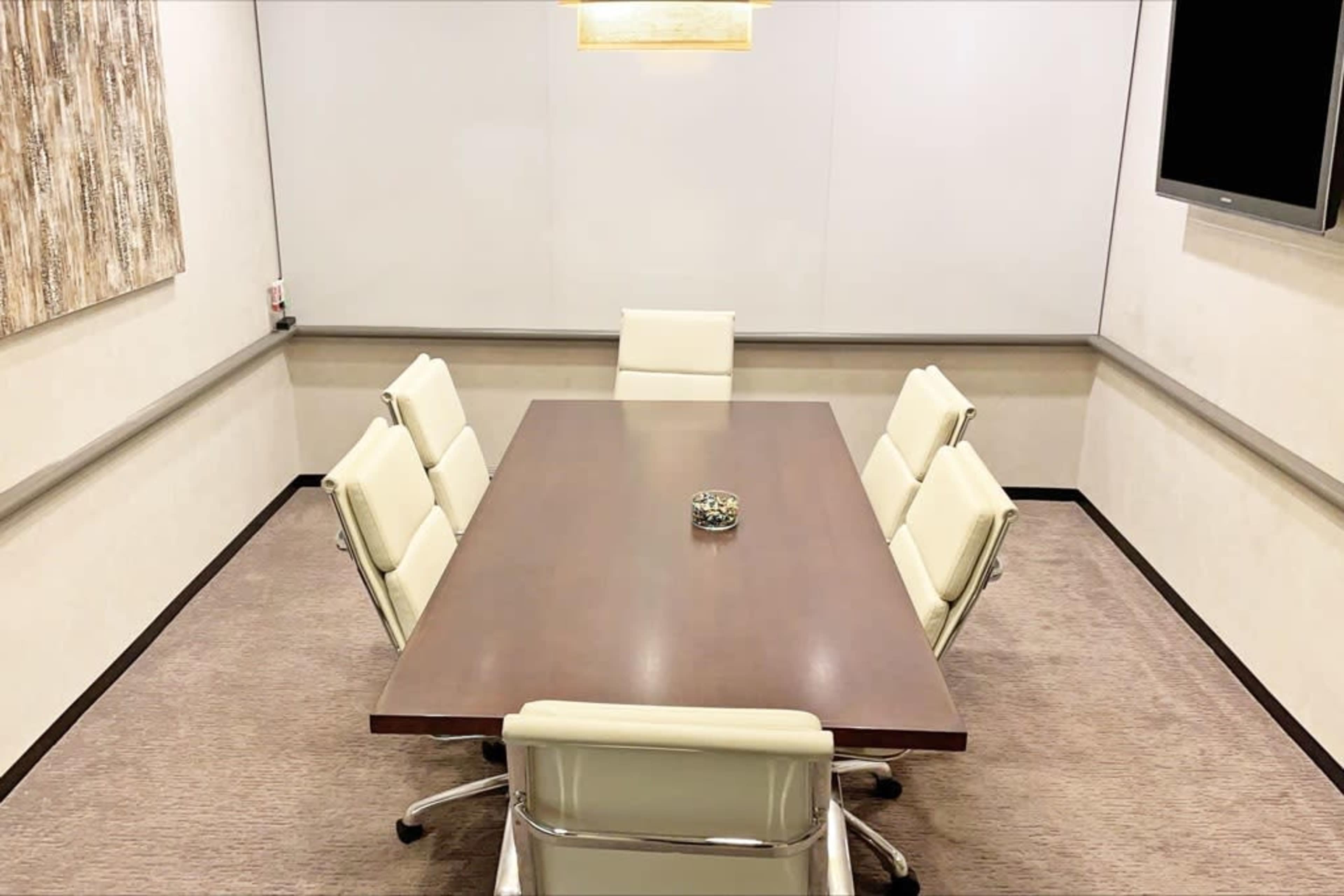 A conference room features a large wooden table surrounded by six white leather chairs, with a decorative piece in the center and blank walls.
