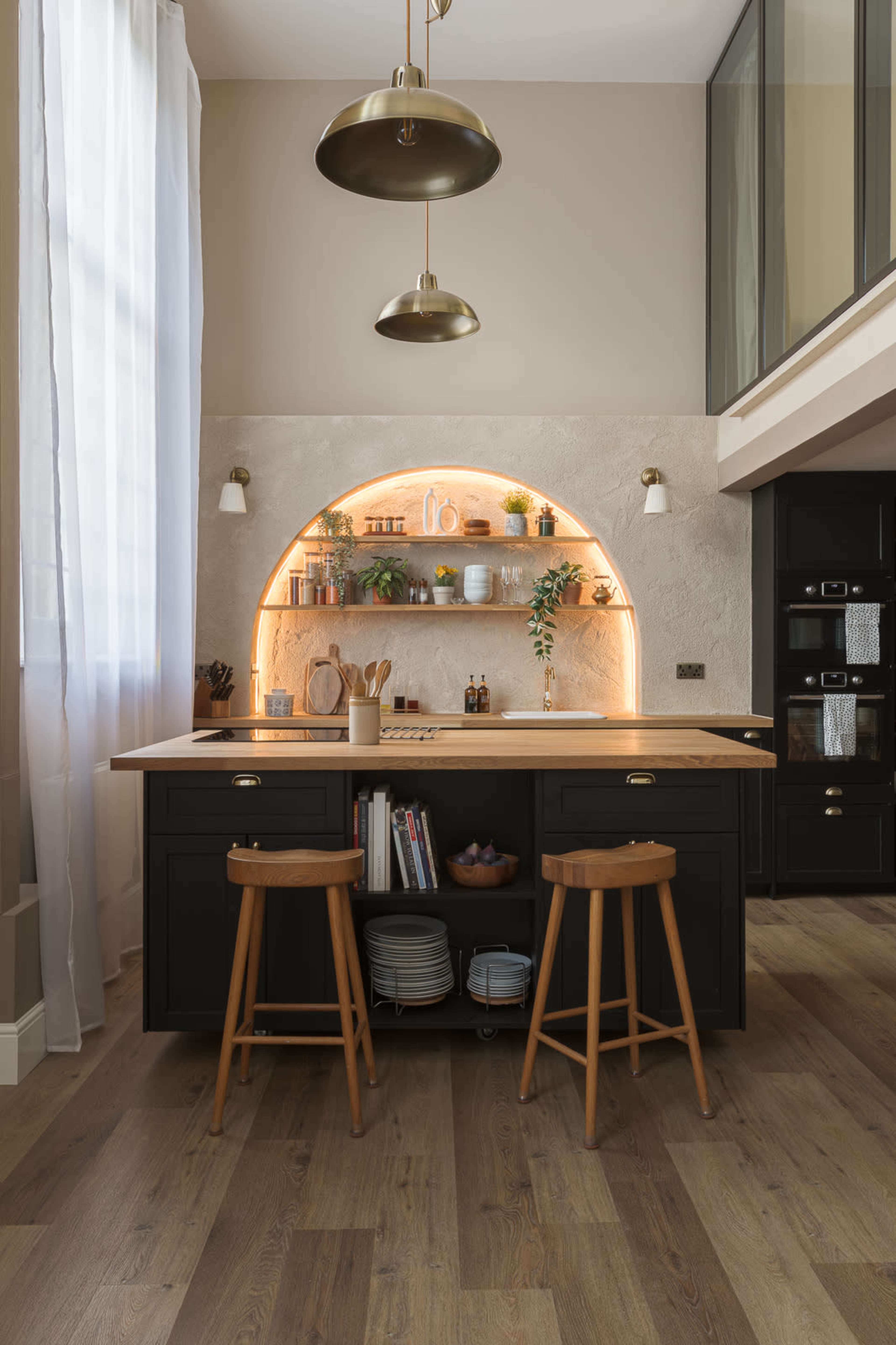 A modern kitchen features a central island with wooden stools, illuminated shelves displaying kitchenware, and large windows allowing natural light to enter.