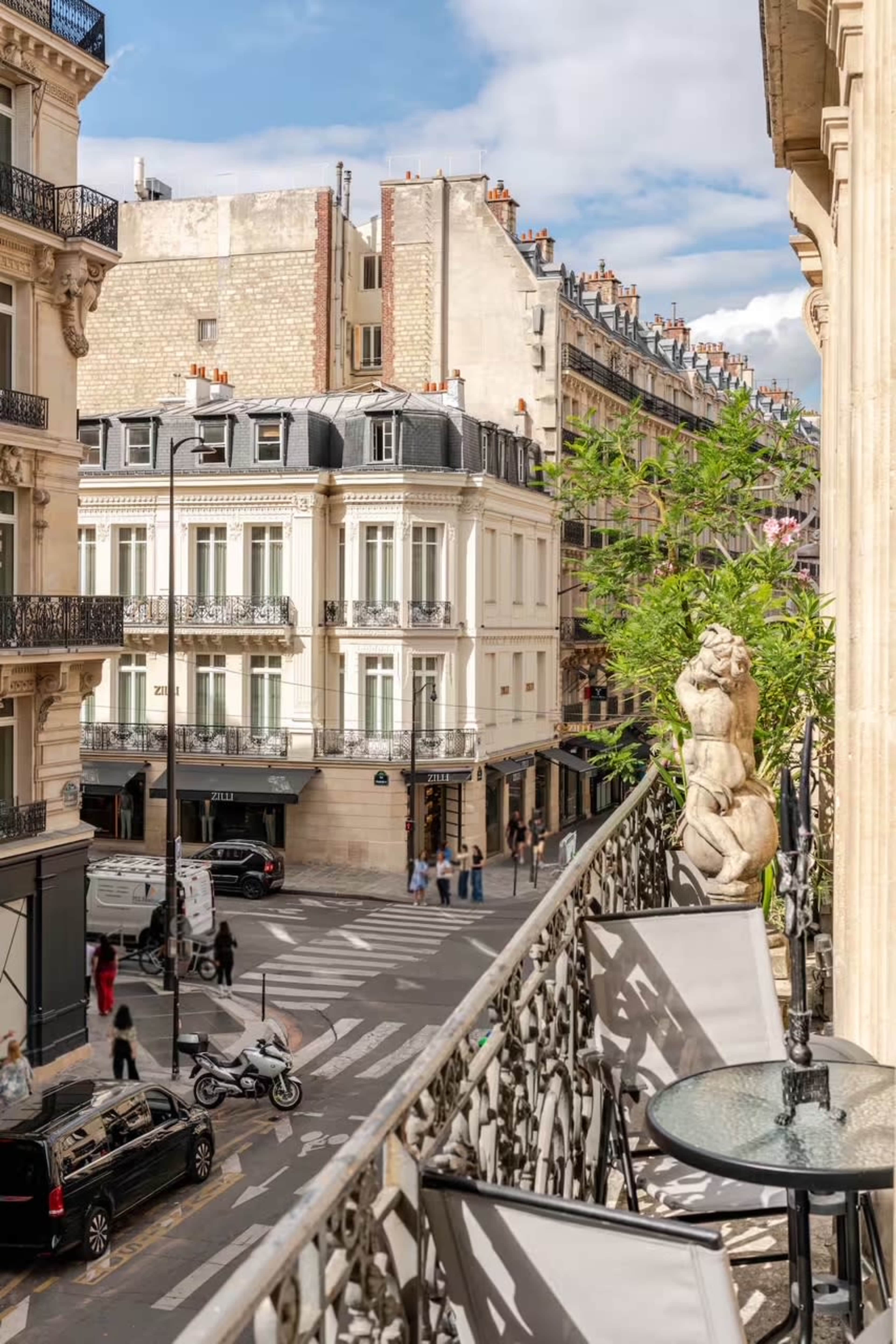 The image shows a Parisian street view from a balcony, featuring elegant buildings, a green plant, and people walking below.