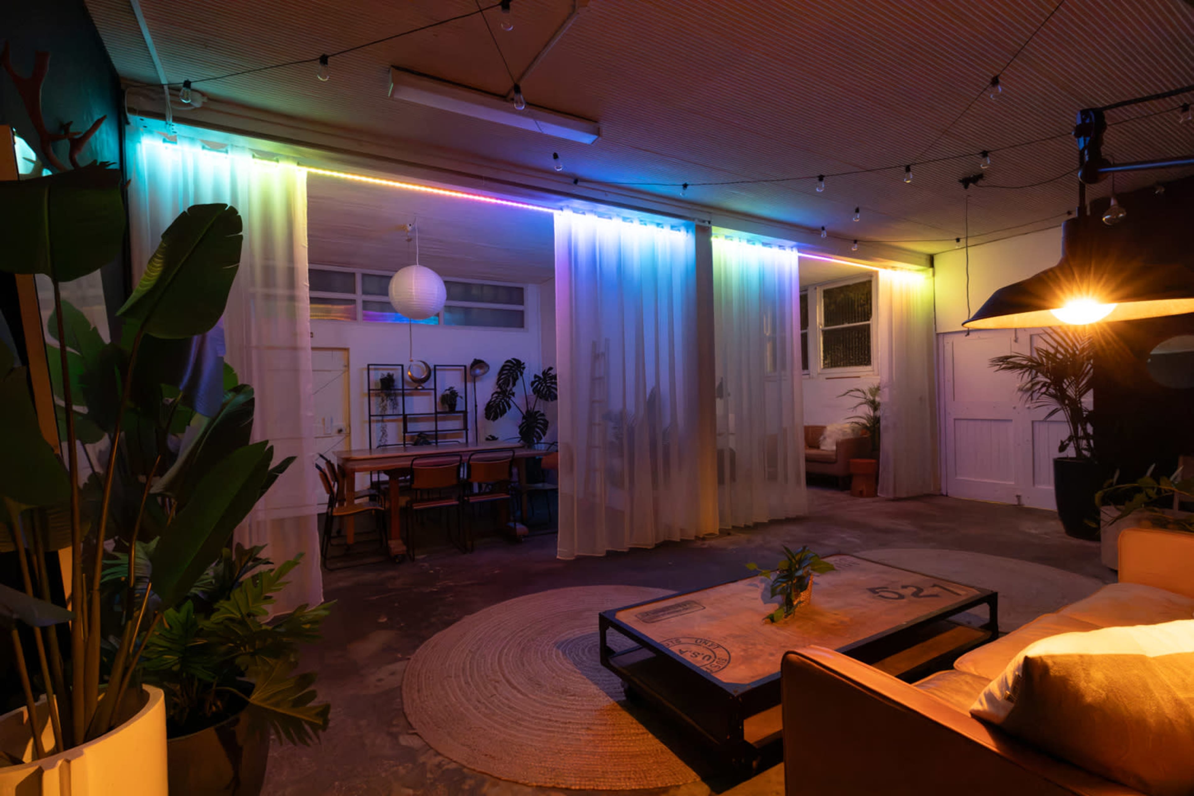 The image shows a spacious indoor living area with soft curtains, decorative plants, and ambient lighting.