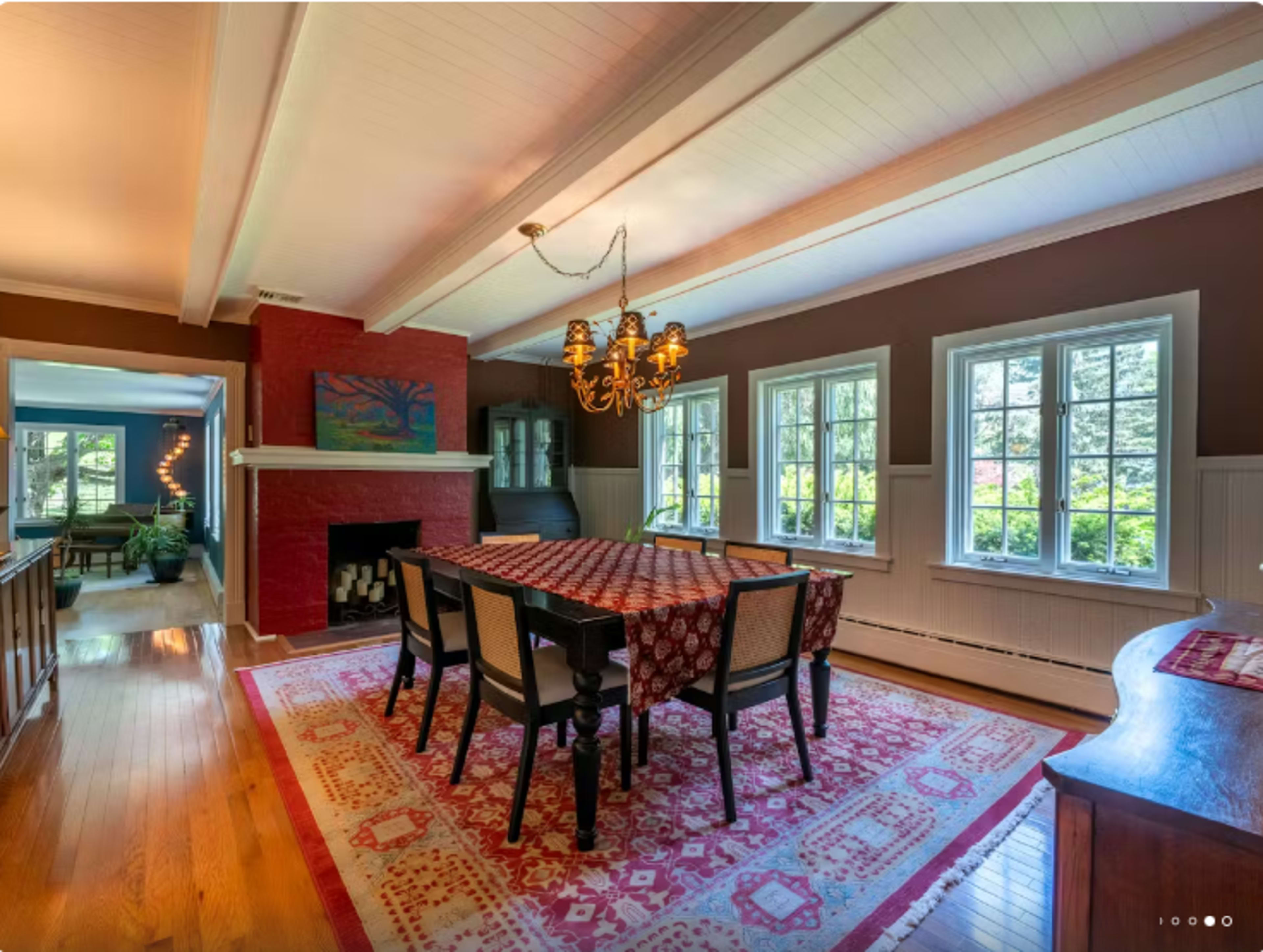 The image shows a dining area with a large table set for meals, surrounded by chairs, and features a red fireplace and multiple windows allowing natural light.
