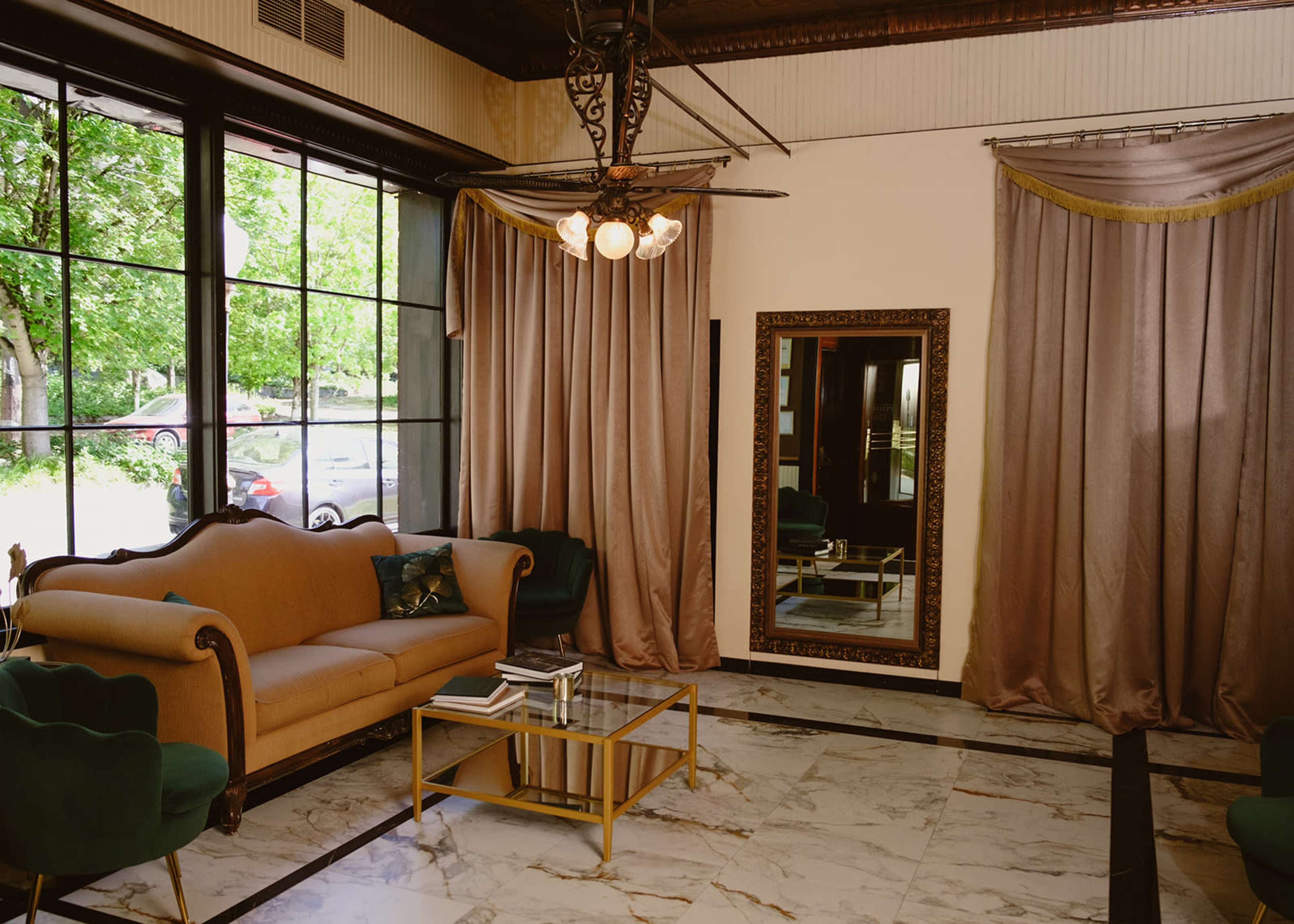 The image shows a stylishly furnished living room with a beige sofa, green accent chairs, a glass coffee table, and large windows draped with curtains.