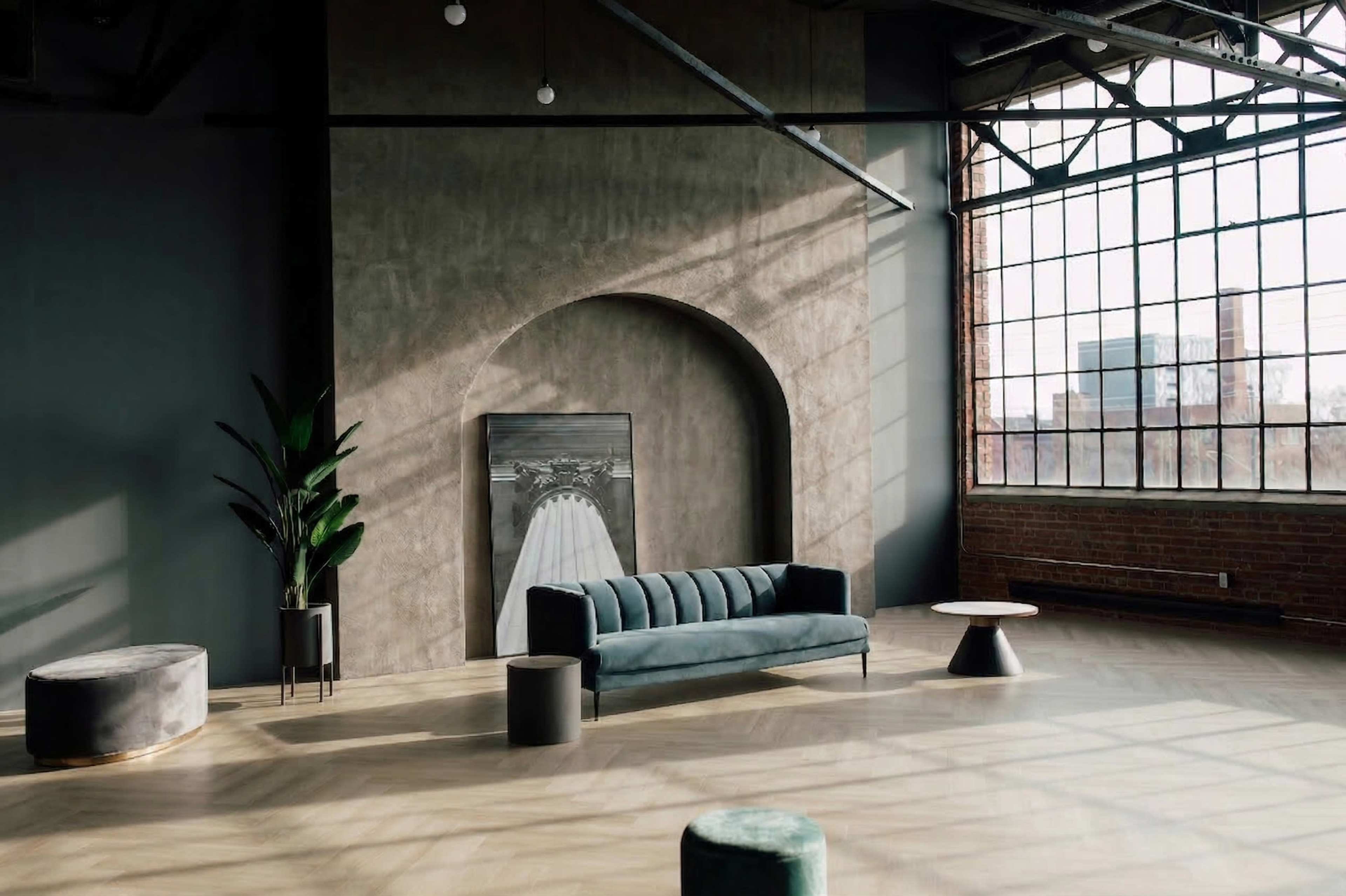 The image features a spacious, modern interior with a velvet sofa, a plant, circular side tables, and large windows allowing natural light to flood the room.