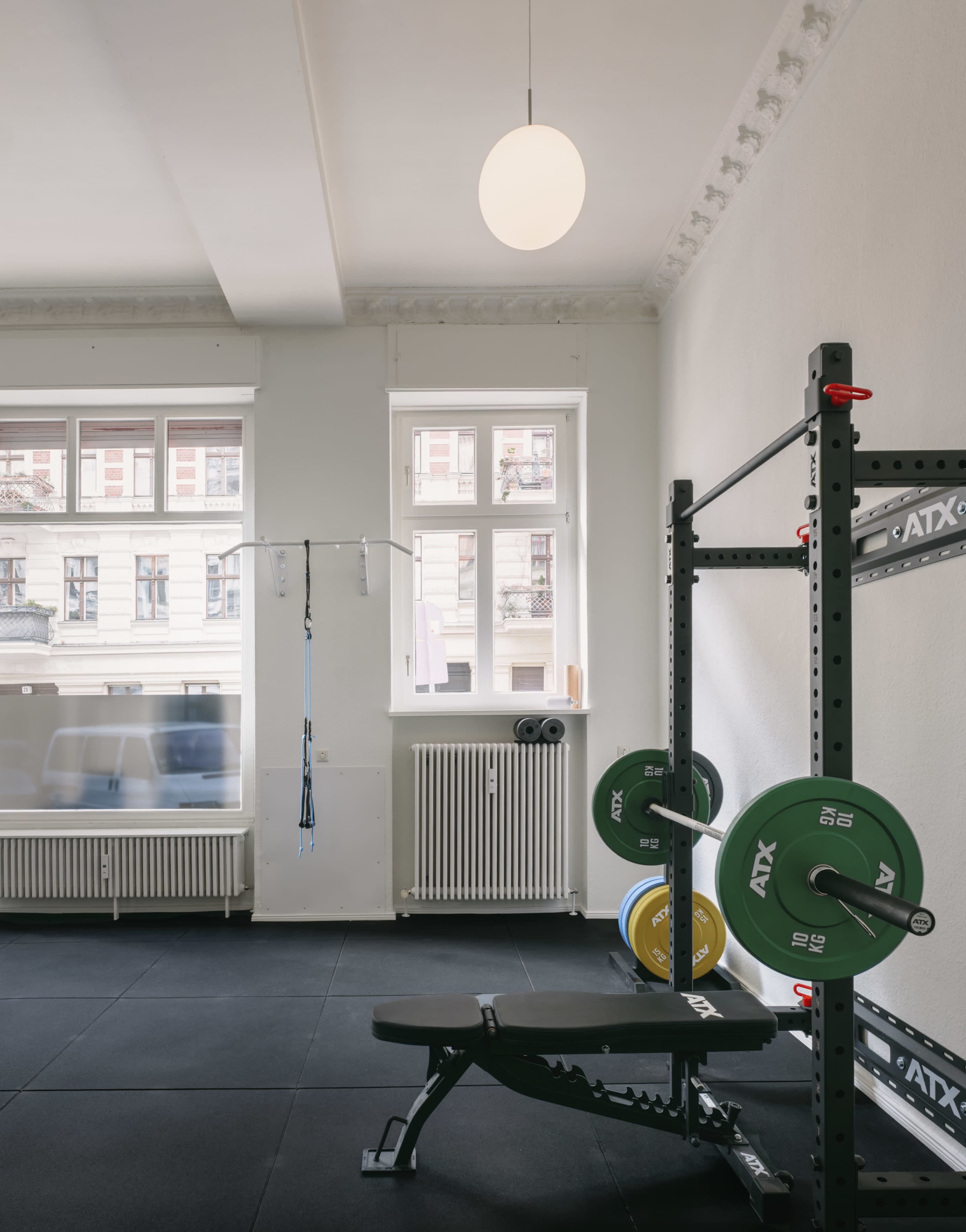 The image shows a bright gym interior featuring a weightlifting rack with barbells, a bench, and workout equipment against a backdrop of large windows.