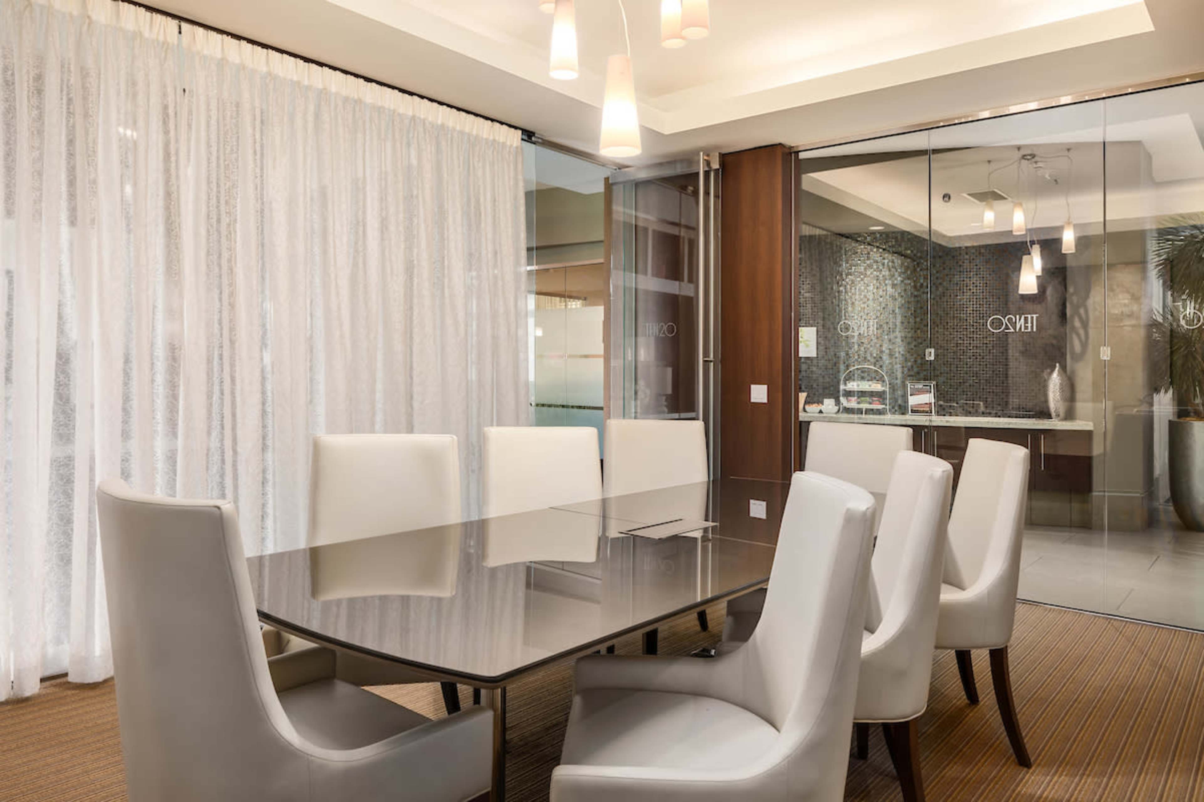 A modern conference room features a glass table surrounded by white chairs and a backdrop of sheer curtains and a glass wall leading to another space.