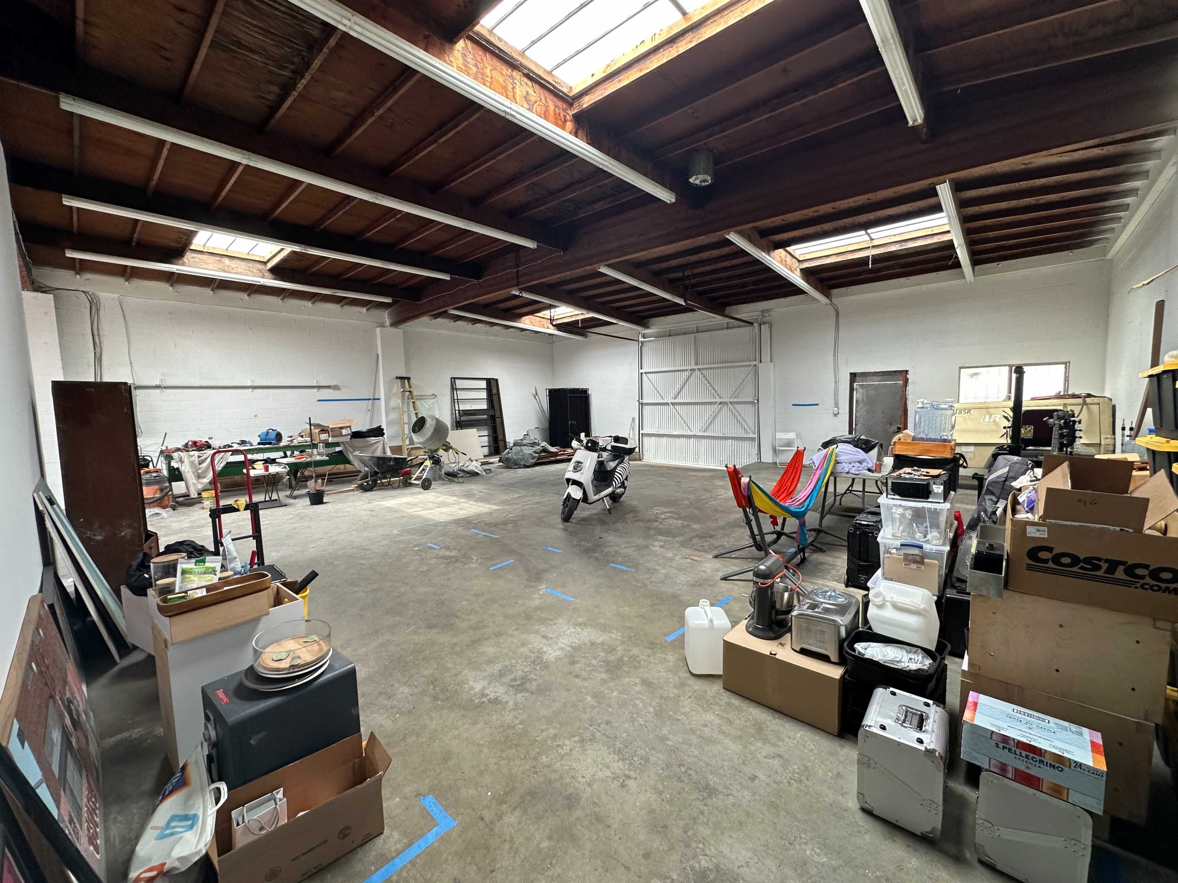 The image shows a spacious, cluttered garage or workshop area filled with various items, including boxes, tools, and a scooter, under a wooden ceiling with skylights.
