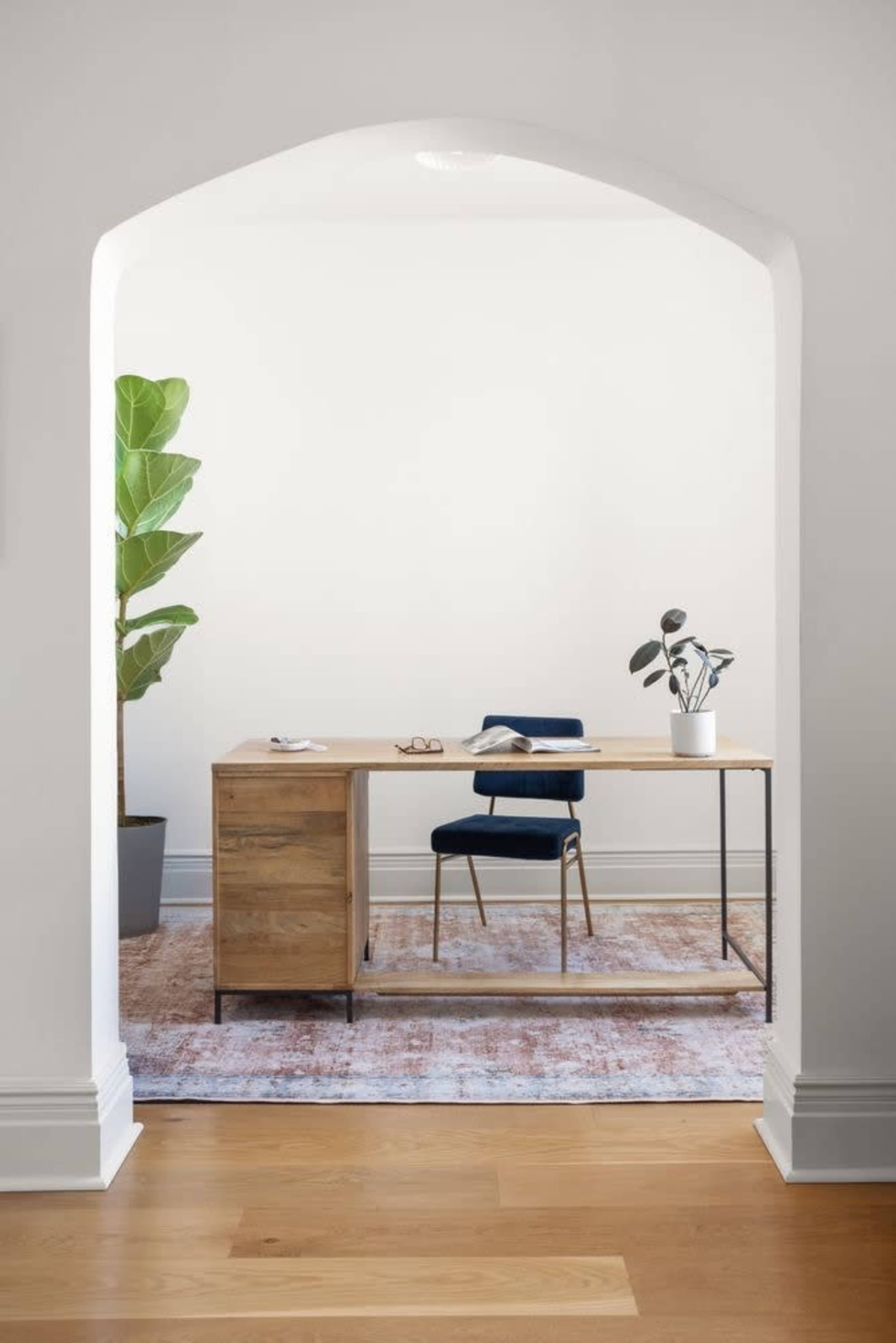 A wooden desk with a chair and potted plants is set in a well-lit room, viewed through an arched opening.