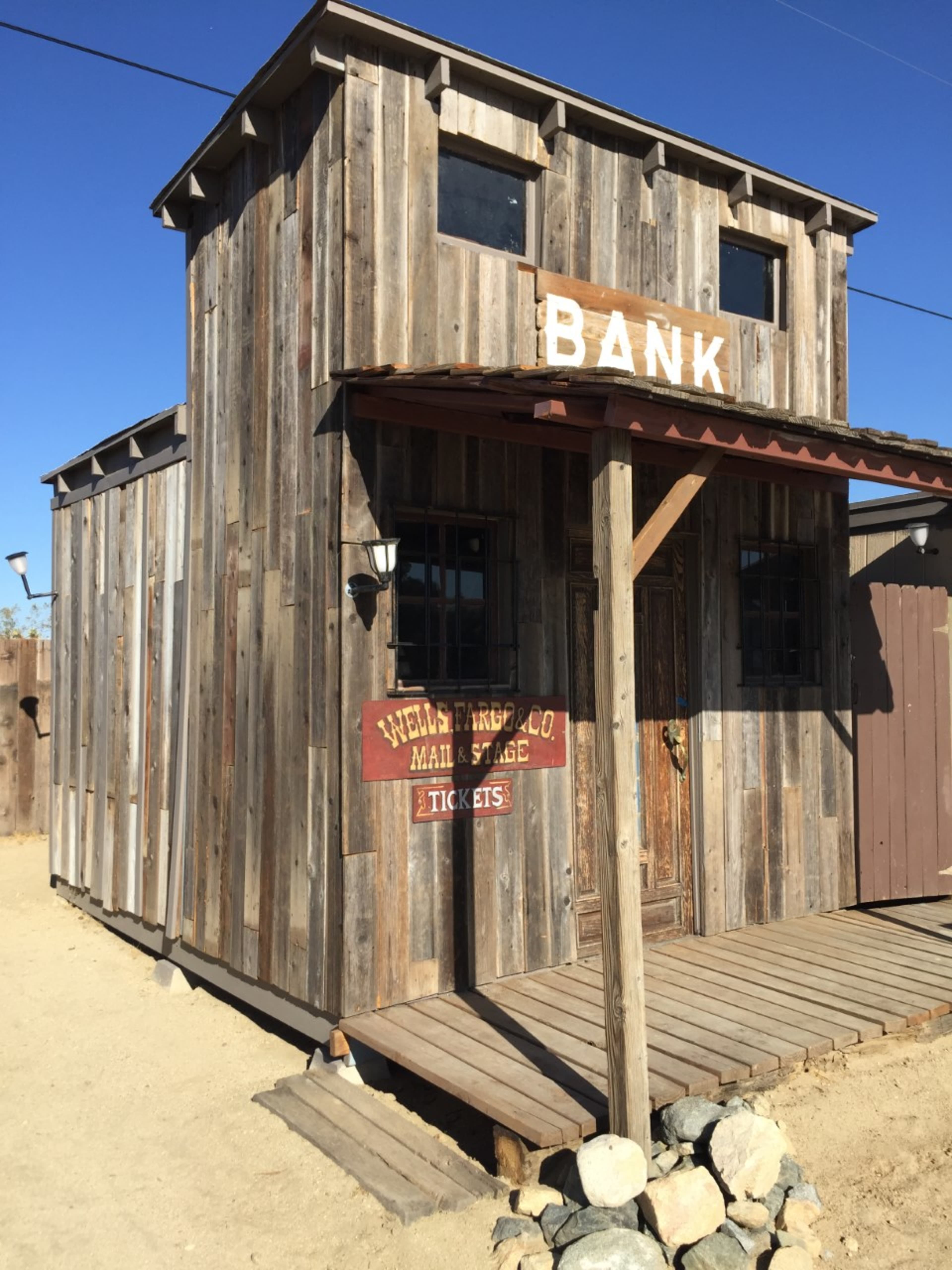 A rustic wooden bank building with a sign that reads "BANK" on the front and an old-fashioned stagecoach ticket sign on the side.
