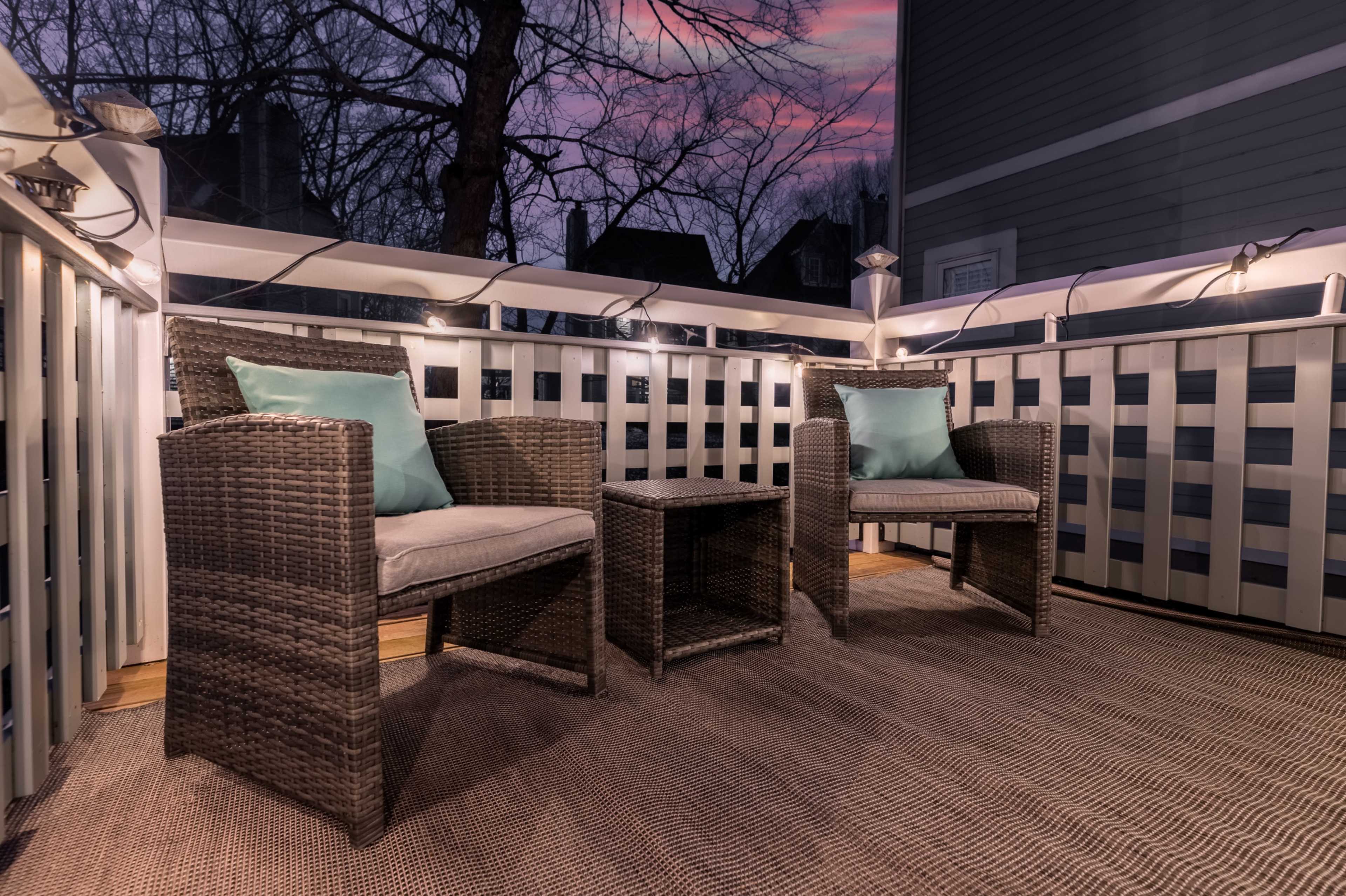 A small balcony features two wicker chairs and a table, surrounded by string lights, with a sunset sky visible in the background.