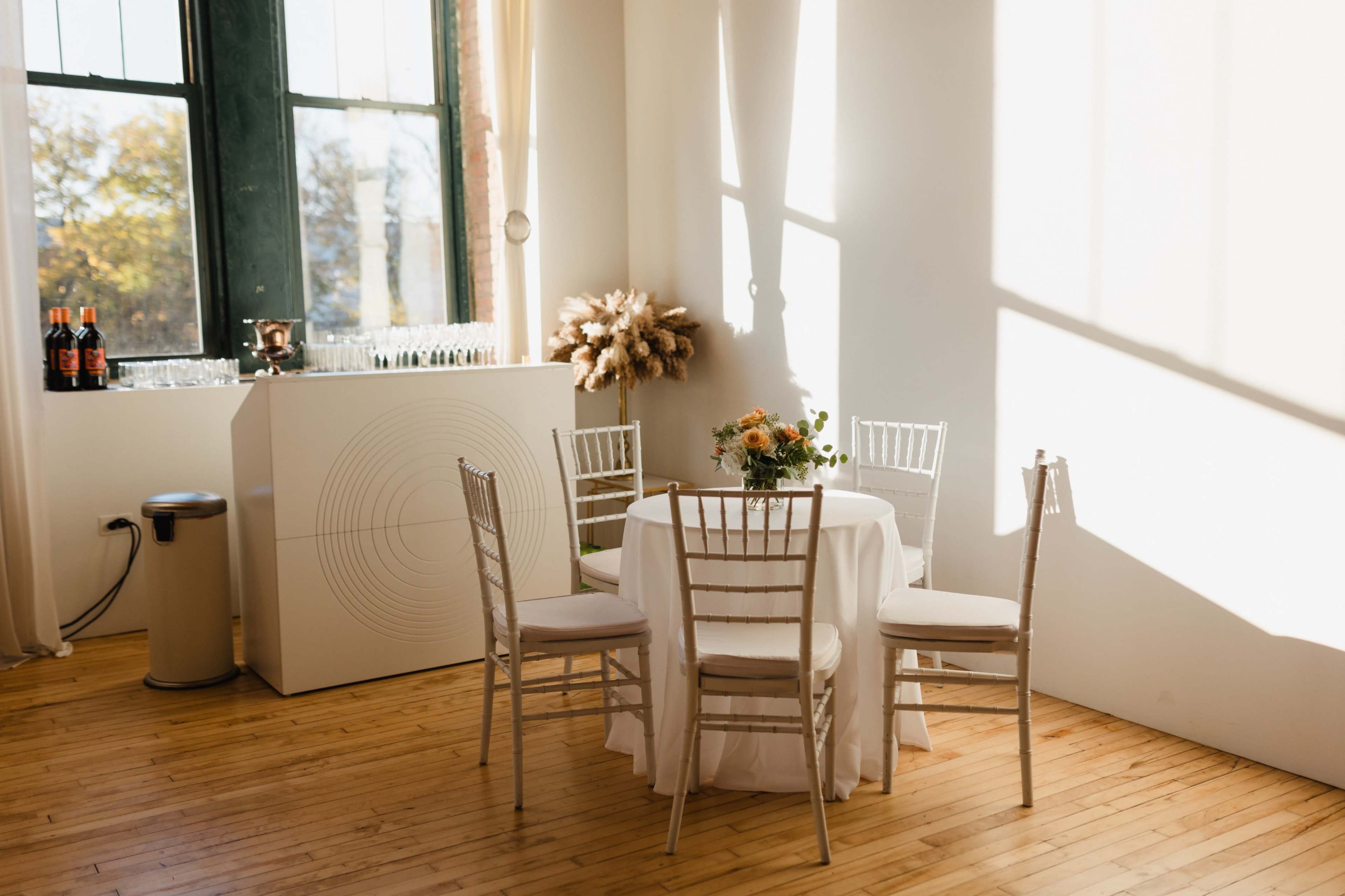 A small round dining table with four chairs is set in a brightly lit room featuring large windows and a bar area in the background.