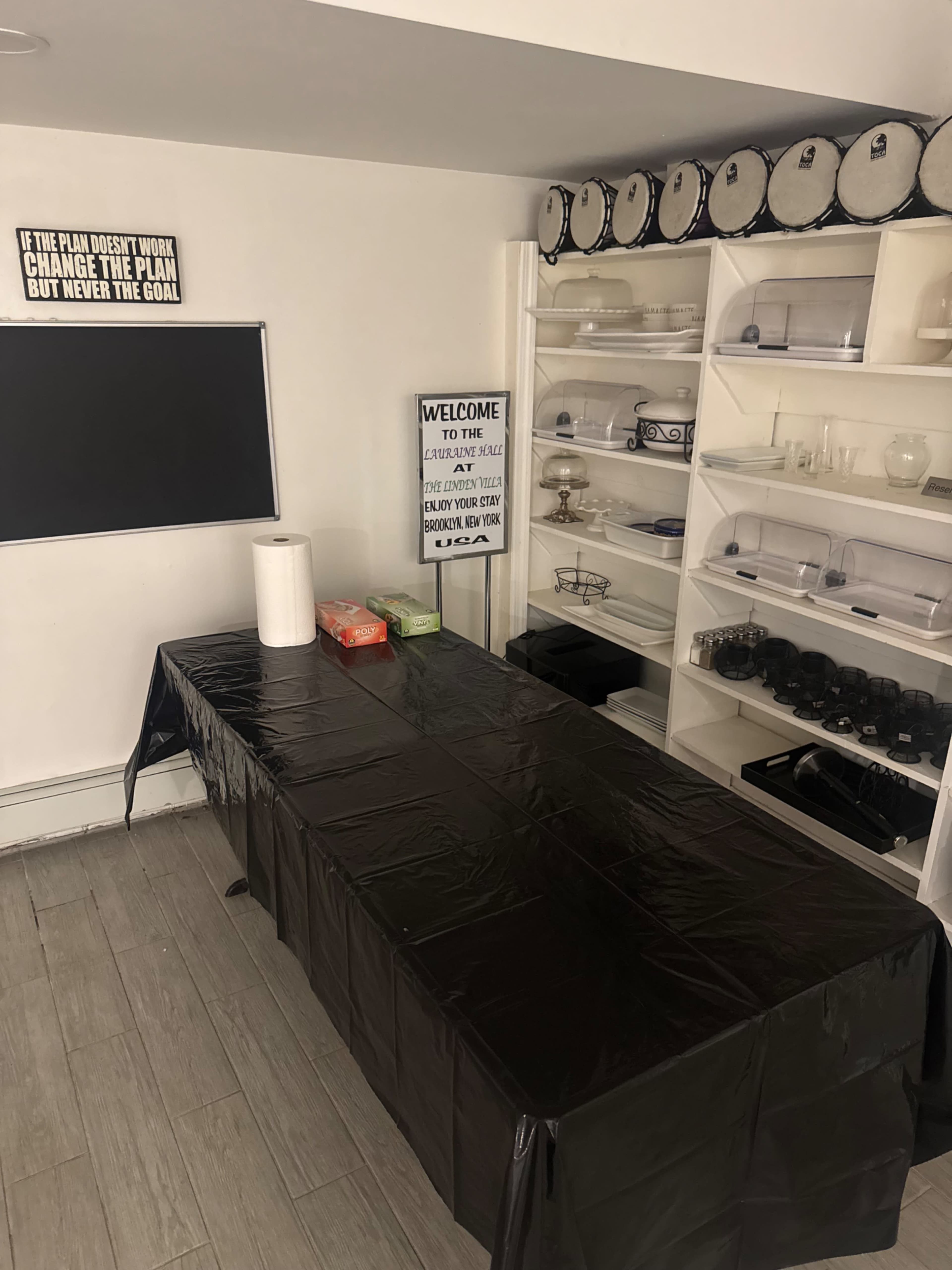 The image shows a large black table covered with a plastic tablecloth in a room with shelves displaying various containers and a wall-mounted screen.