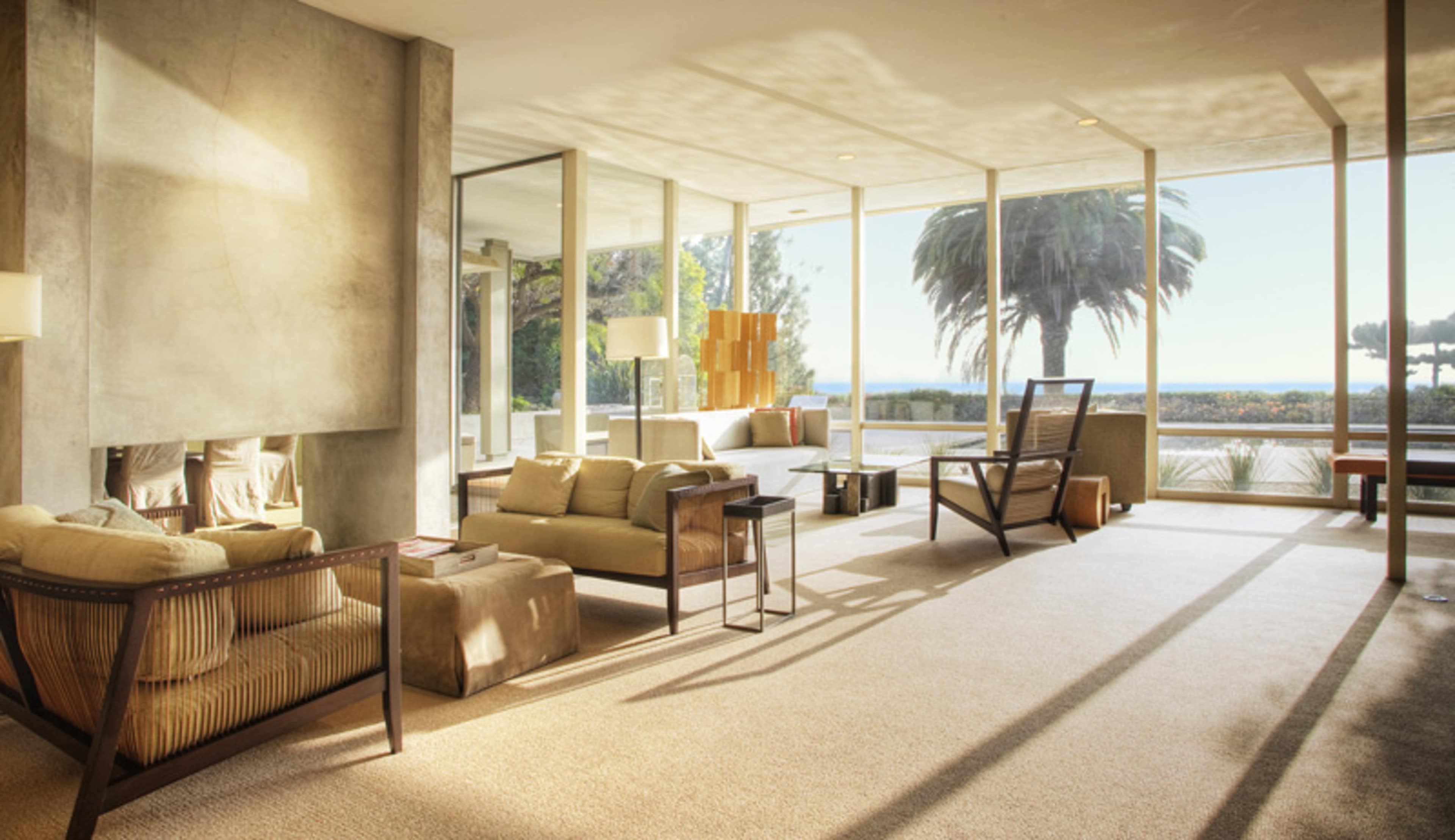 A modern living room features large windows that overlook a palm tree and outdoor scenery, with stylish furniture arranged for comfort.