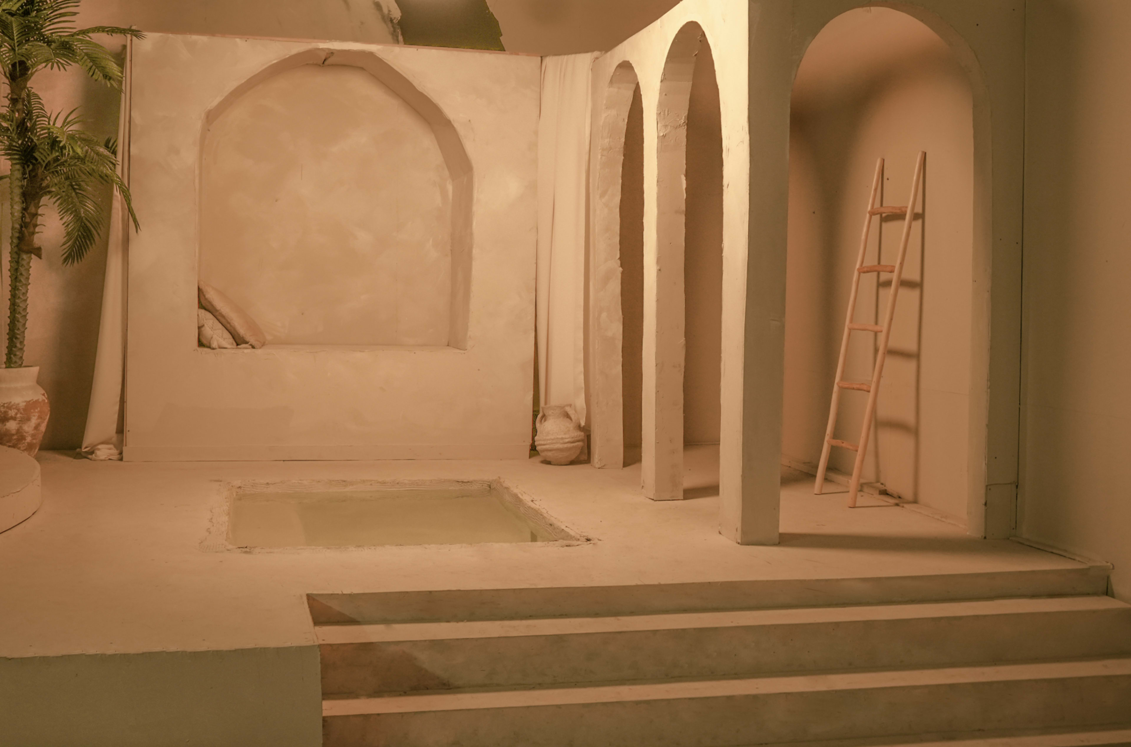 The image shows a minimalist interior scene with arched structures, a small pool, and a ladder against a light-colored wall.