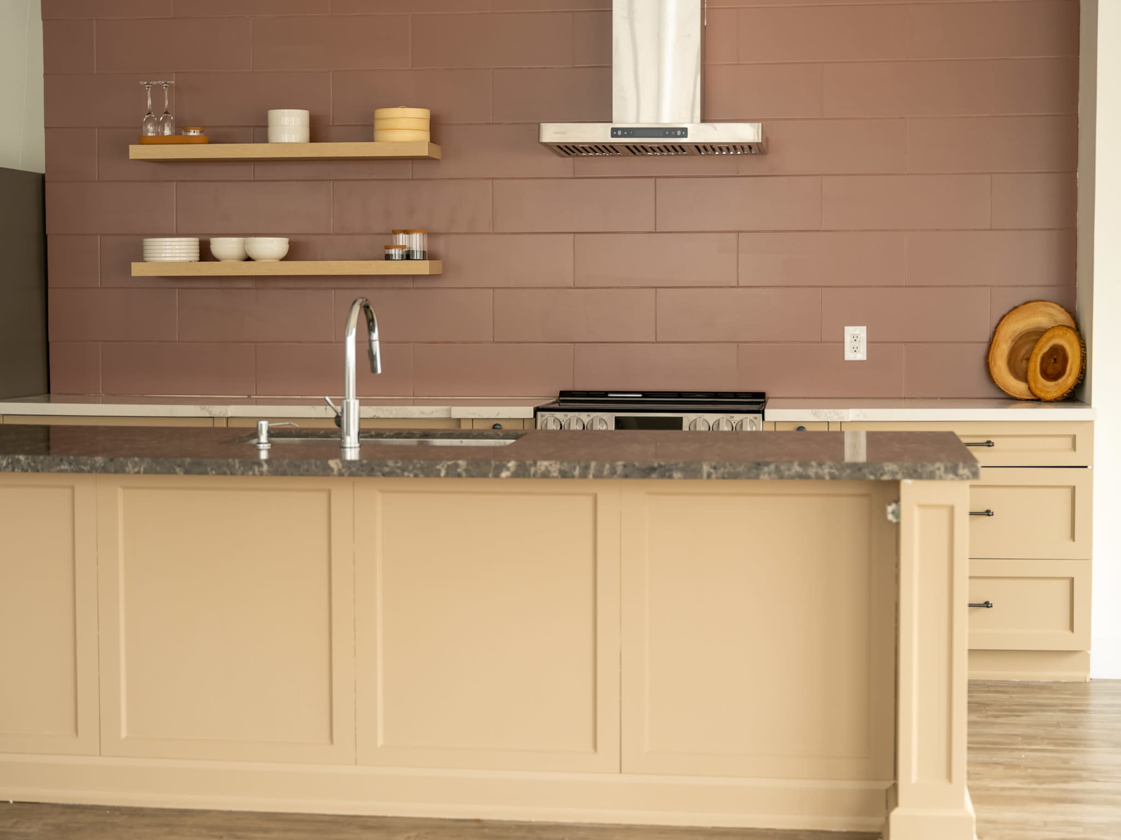 The image shows a modern kitchen with a beige island, a purple accent wall, open shelving, and a stovetop under a metallic hood.
