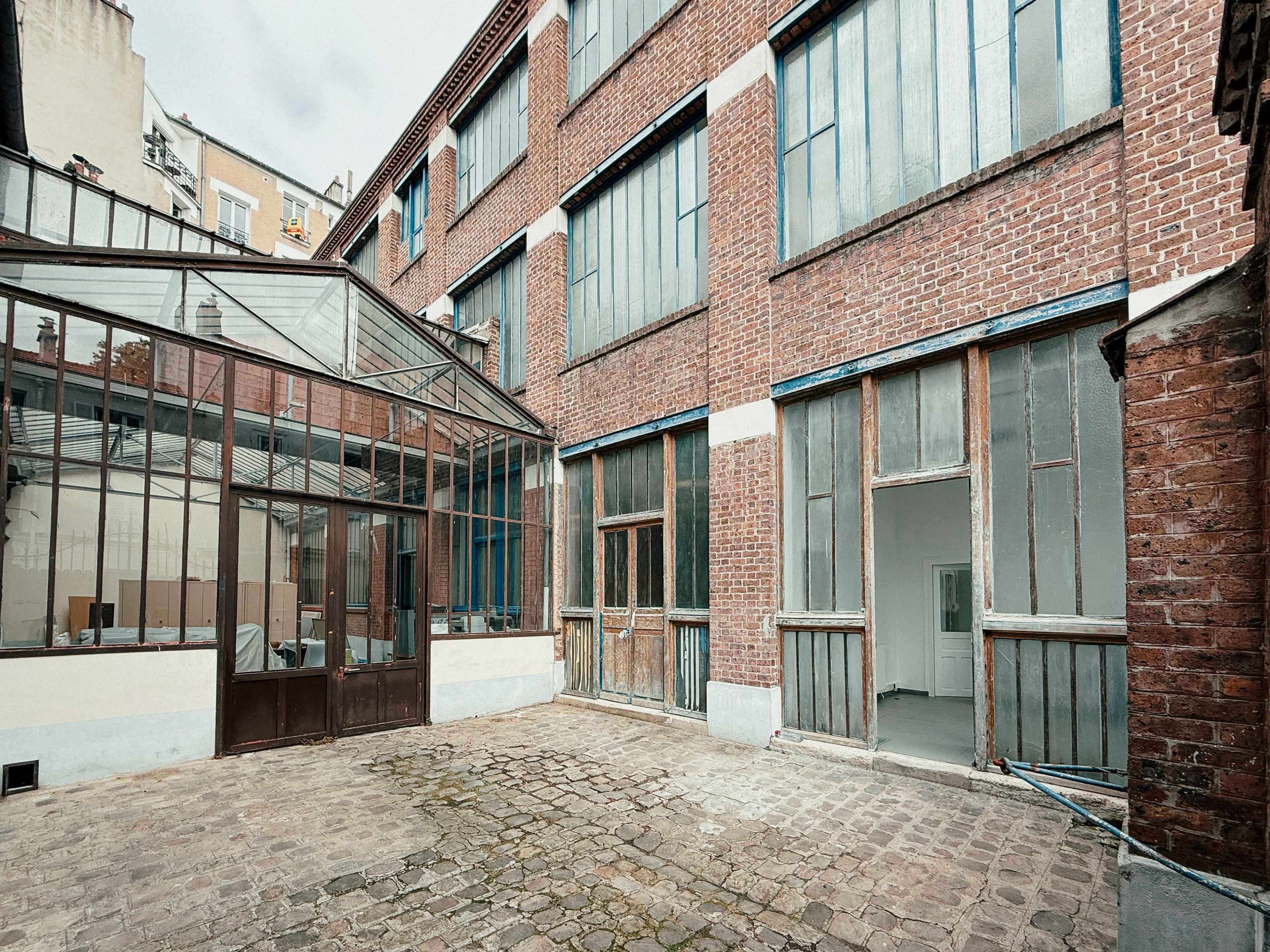 The image shows a courtyard with a brick building featuring large glass windows and a connected greenhouse structure.