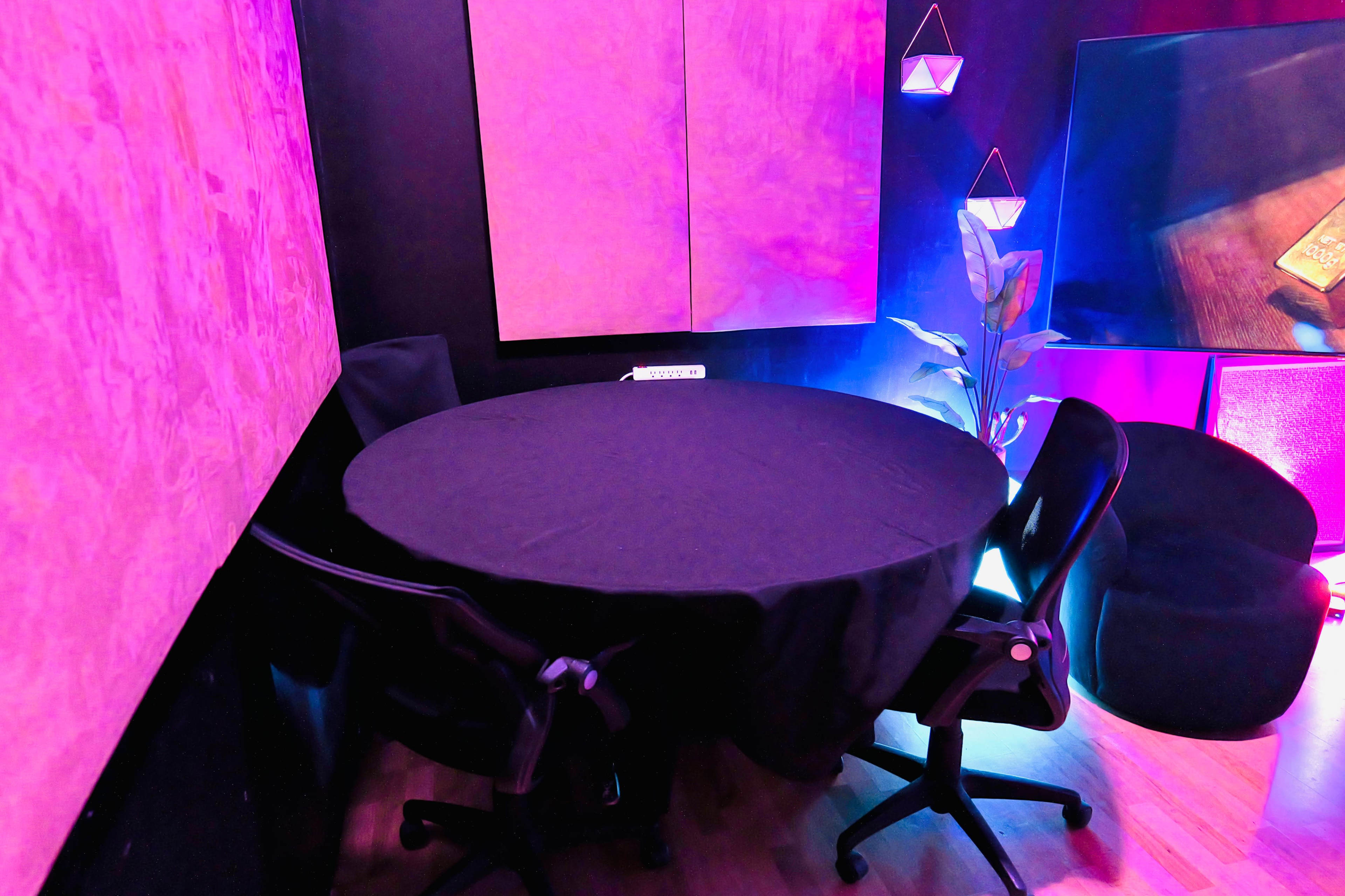 A small room featuring a round black table surrounded by two chairs, illuminated by pink lighting.