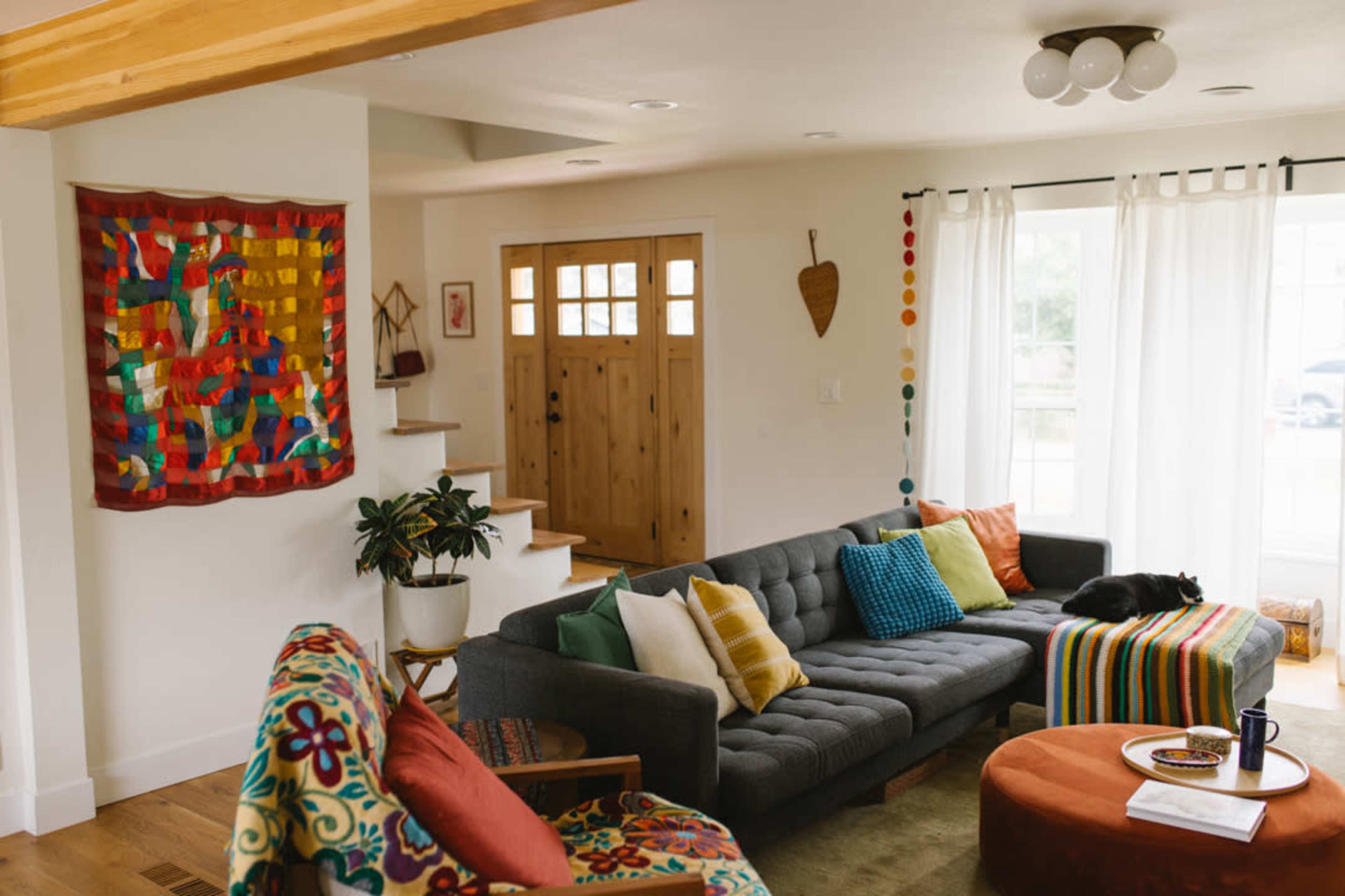 A brightly decorated living room features a gray sofa with colorful pillows, a round orange table, and a staircase leading to a front door adorned with wooden accents.