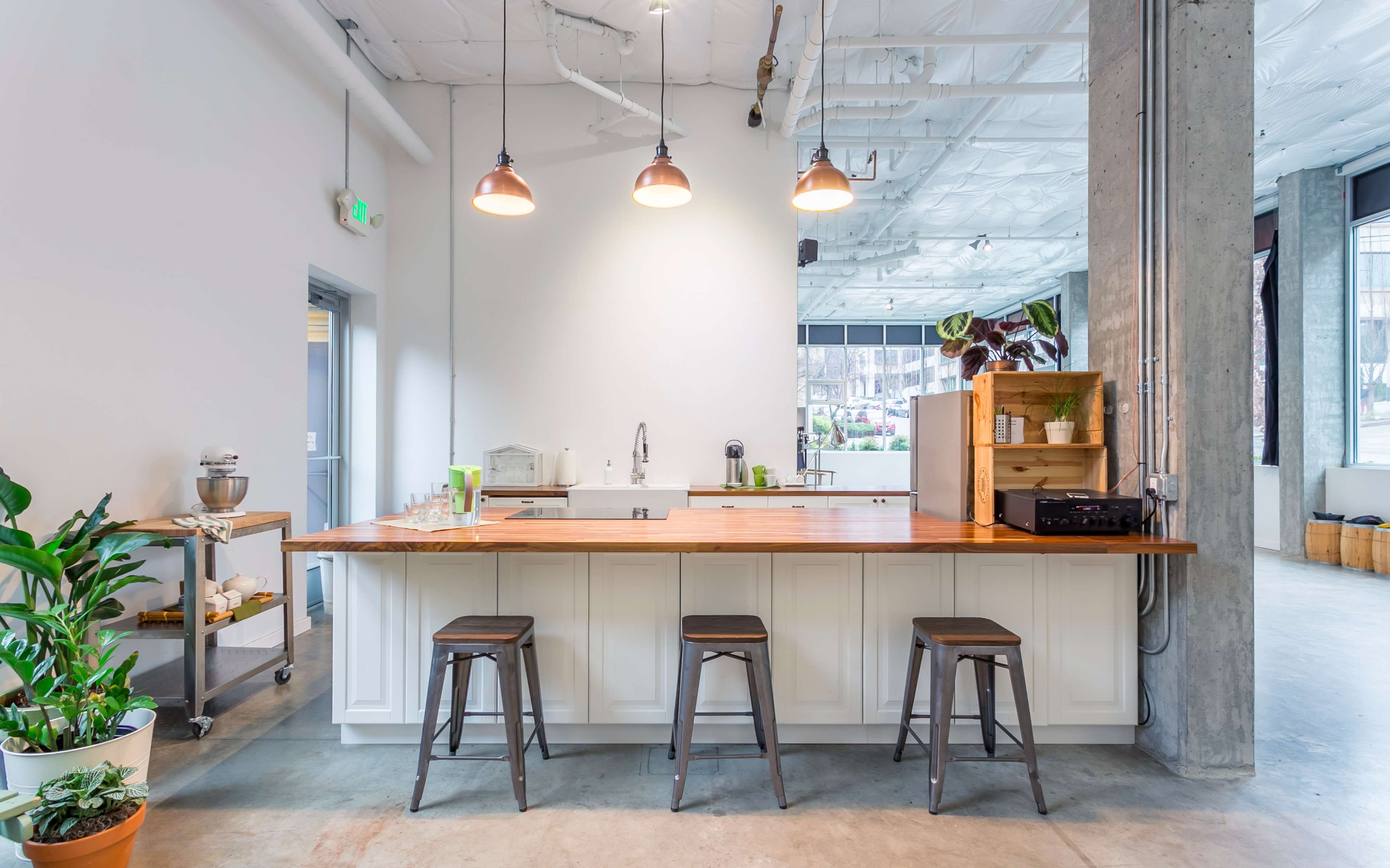A modern kitchen with a wooden bar, three stools, and overhead pendant lights, set in a spacious, well-lit area with large windows and greenery.