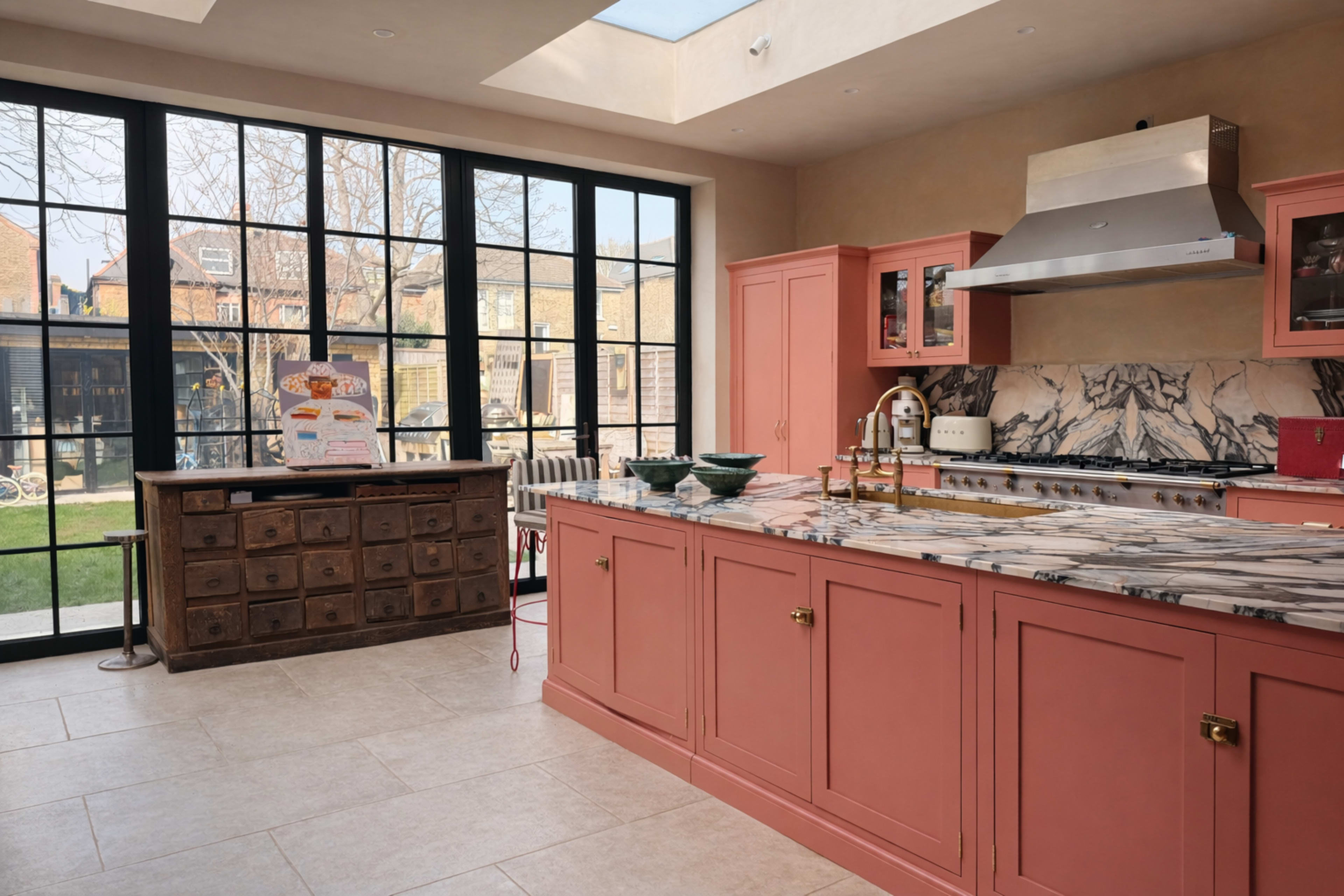 A modern kitchen features a pink cabinetry with a marble countertop, a large window overlooking a garden, and a wooden storage chest against the wall.
