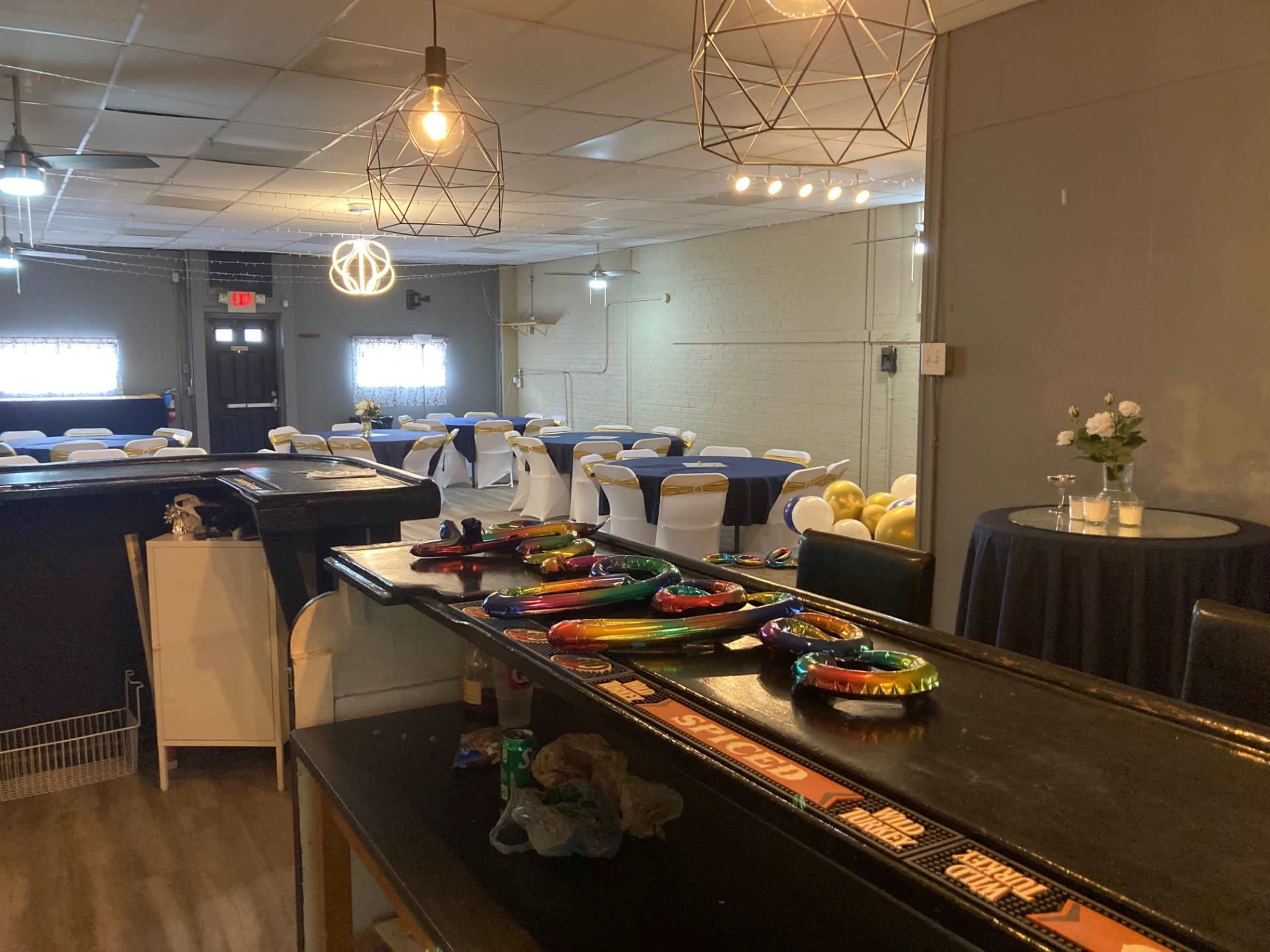 The image shows a spacious event venue with decorated tables and chairs, a bar area in the foreground, and pendant lights illuminating the setting.
