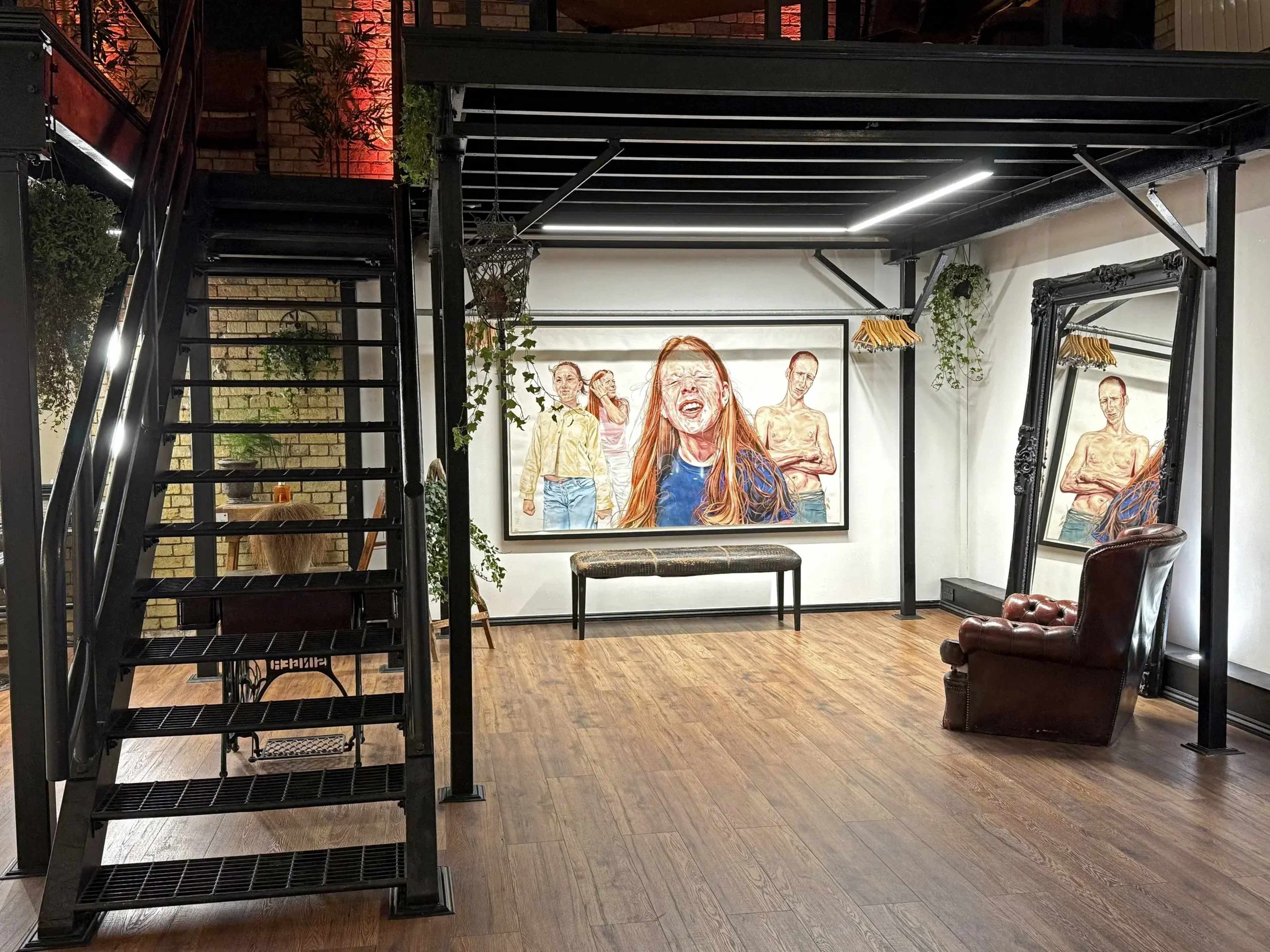 The image shows an industrial-style interior with a metal staircase, a large artwork on the wall featuring people, a wooden bench, and a leather armchair.