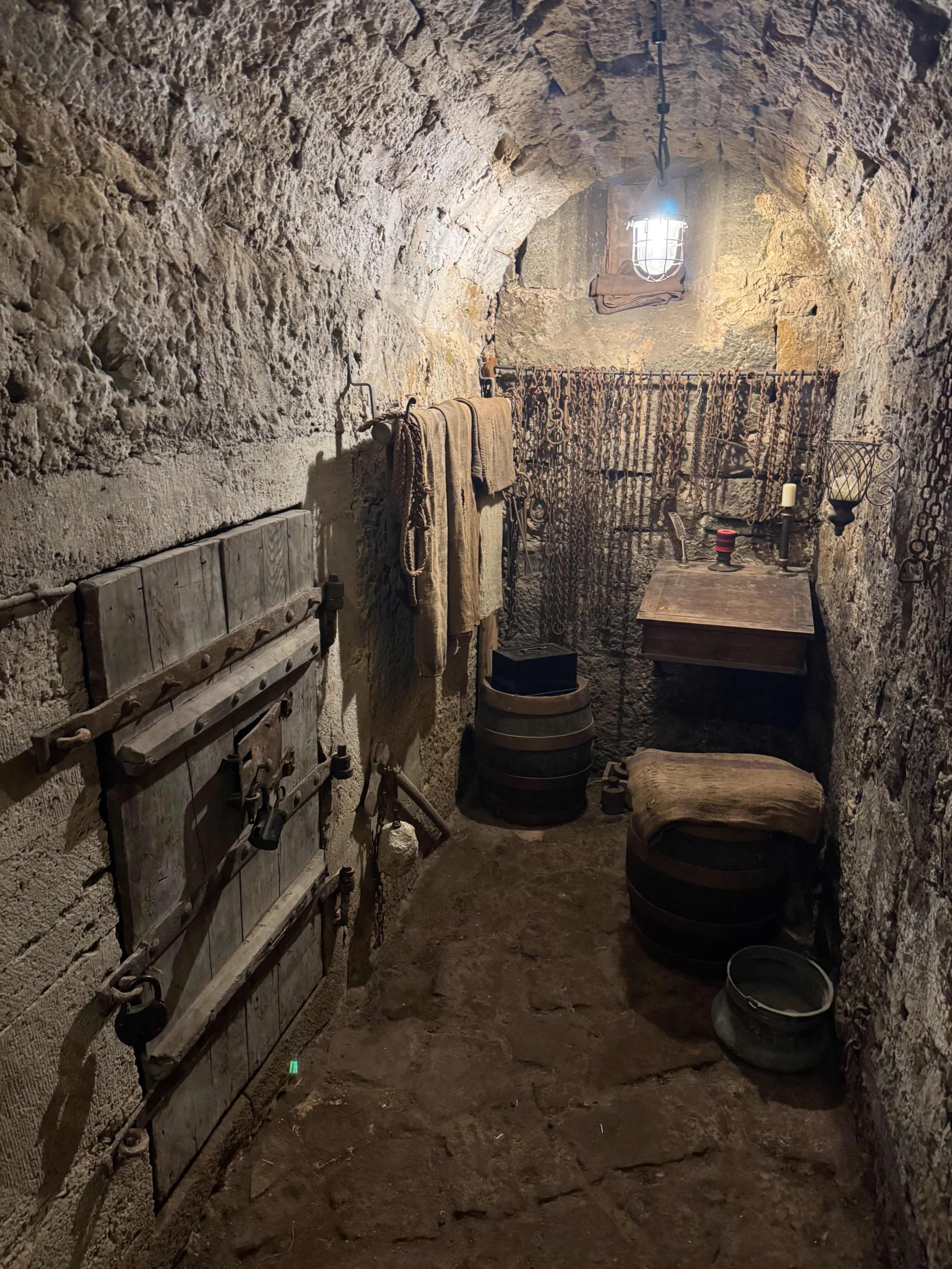 The image shows a stone-walled cellar with wooden barrels, a table, and hanging cloths, illuminated by a lantern.