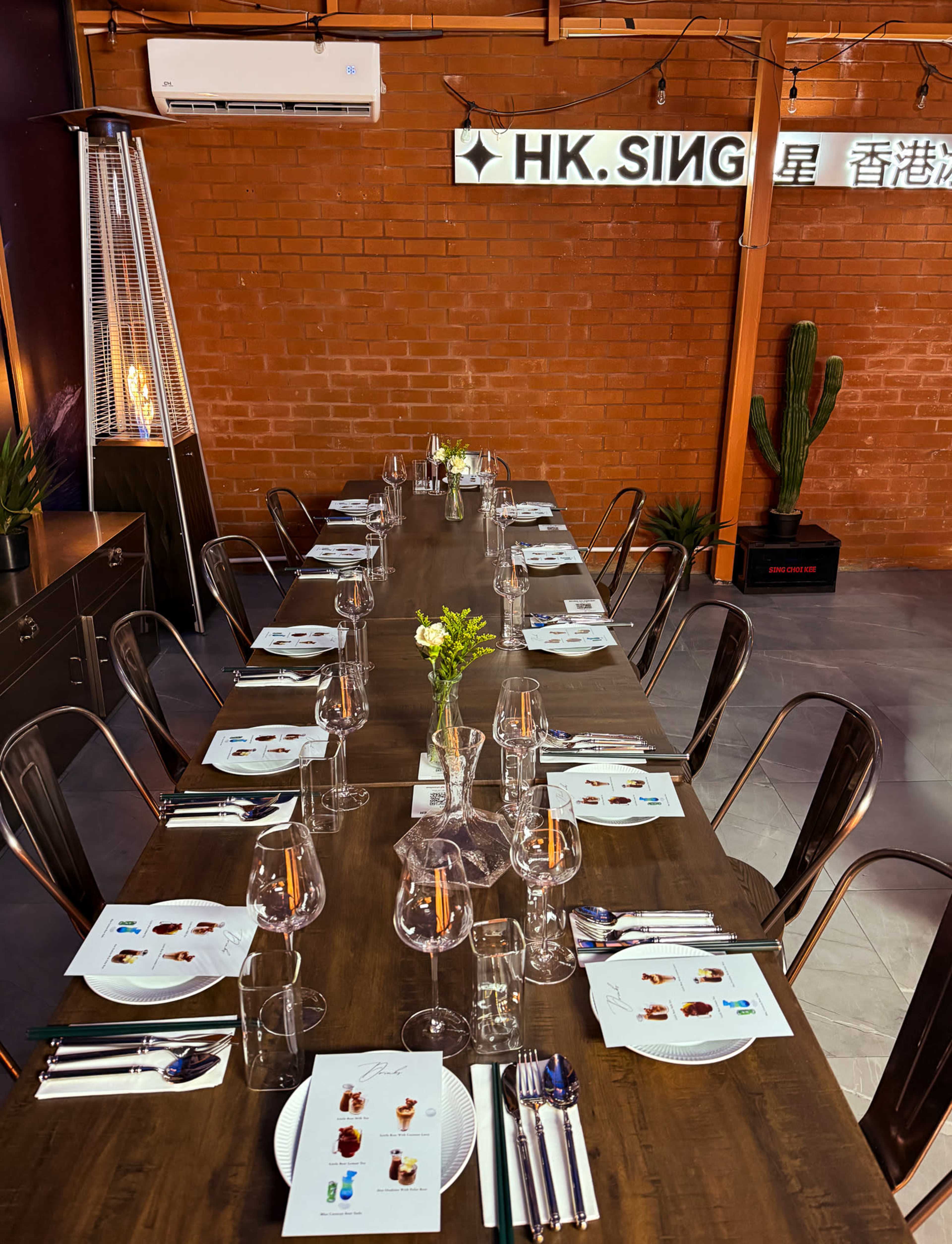 A long wooden dining table is set for a meal, featuring plates, glasses, and utensils, with a small vase of flowers as a centerpiece in a modern restaurant space.