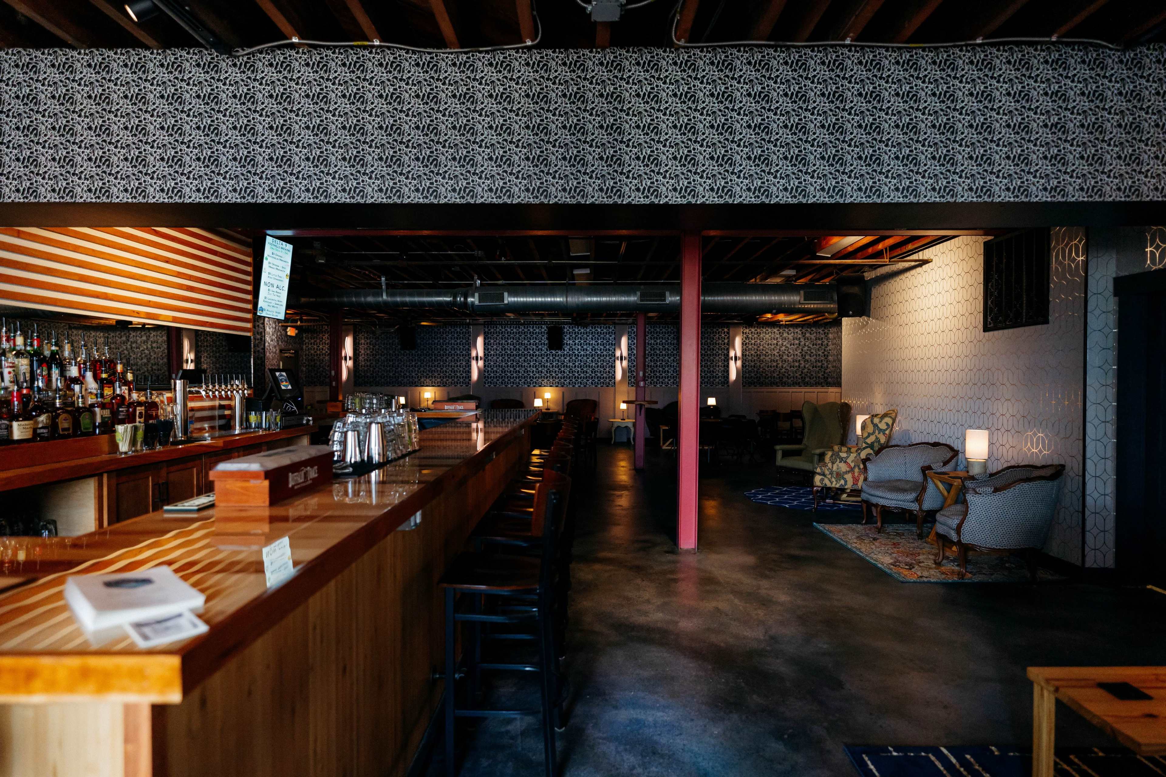 The image shows a nearly empty bar with a long wooden counter, shelves stocked with liquor, and a seating area featuring two armchairs in a dimly lit space.