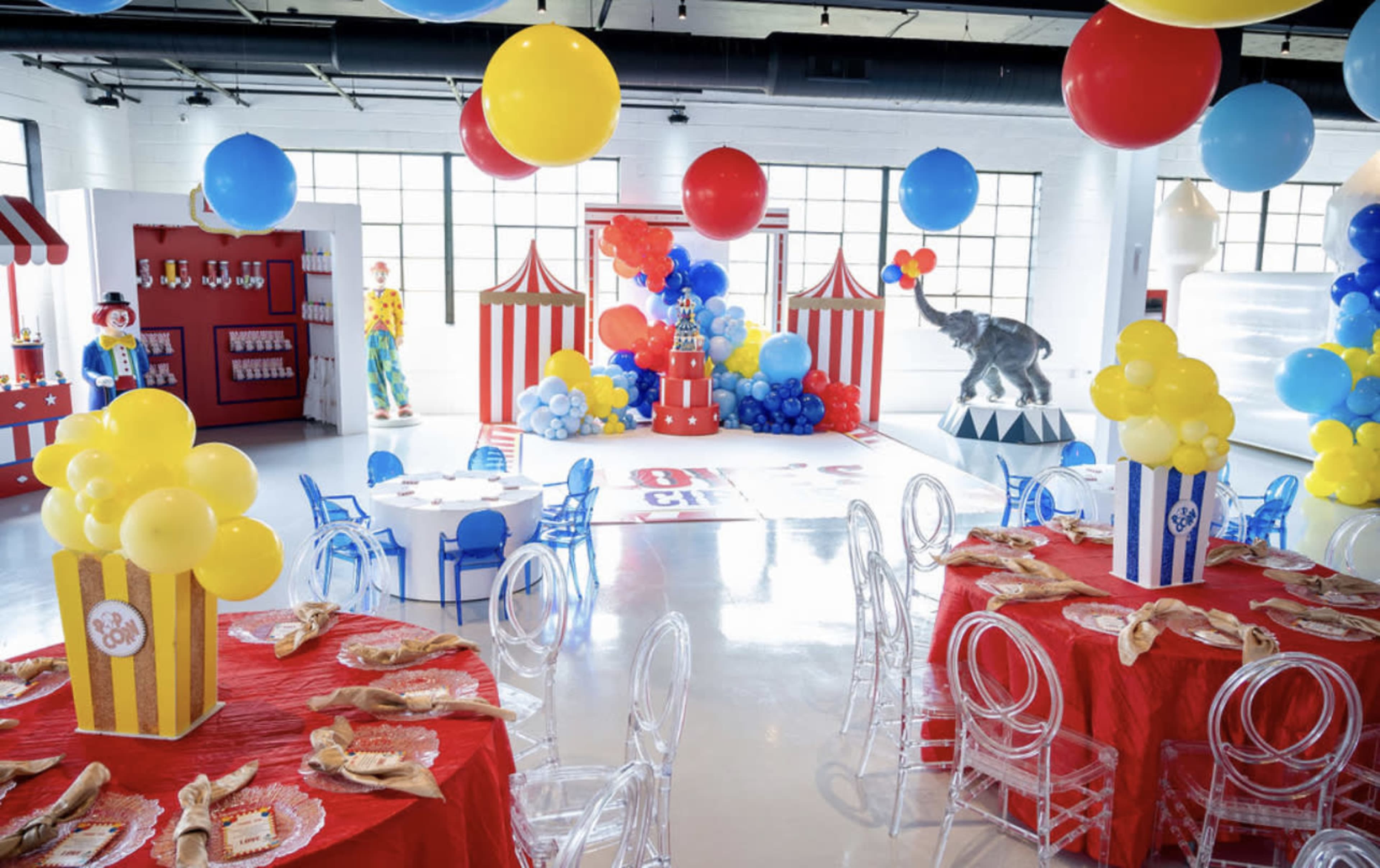 A vibrant carnival-themed party room with colorful tables, balloons, and decorations, including a large elephant figure and clown performers.