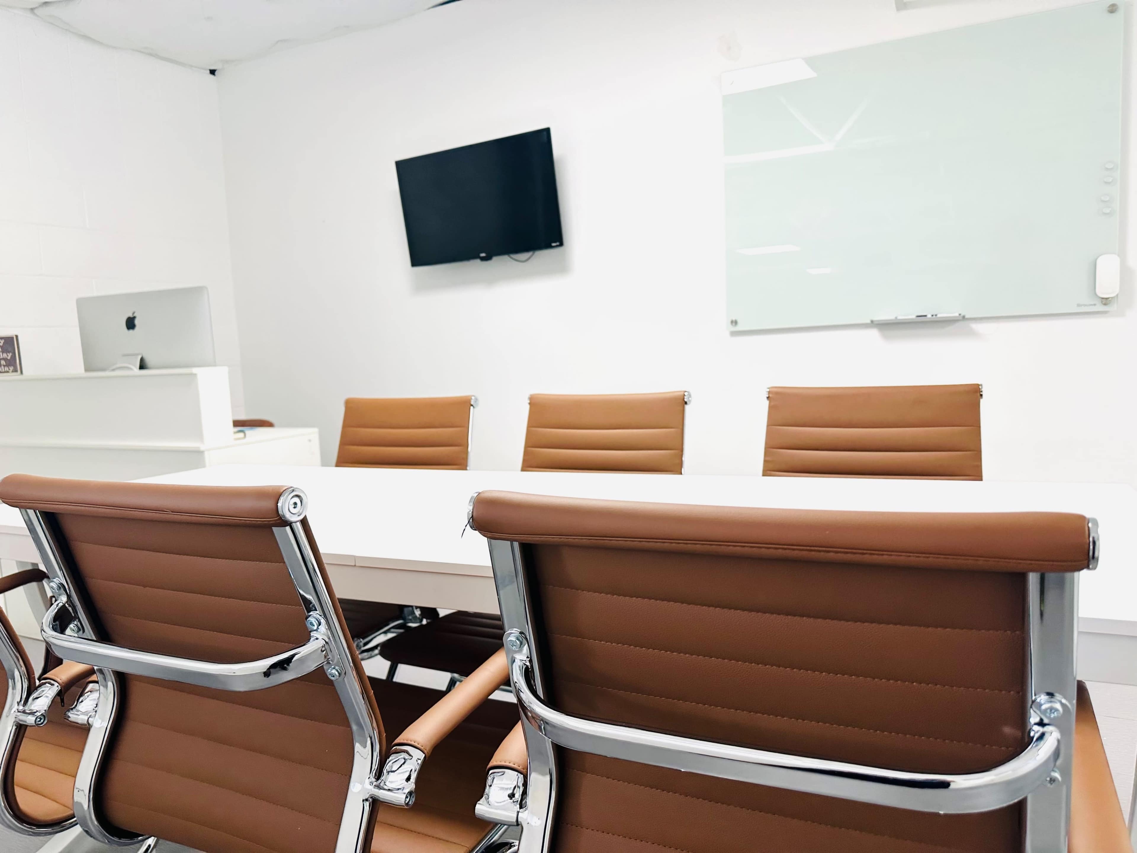 A modern conference room features a long table with four brown leather chairs, a wall-mounted television, and a whiteboard.