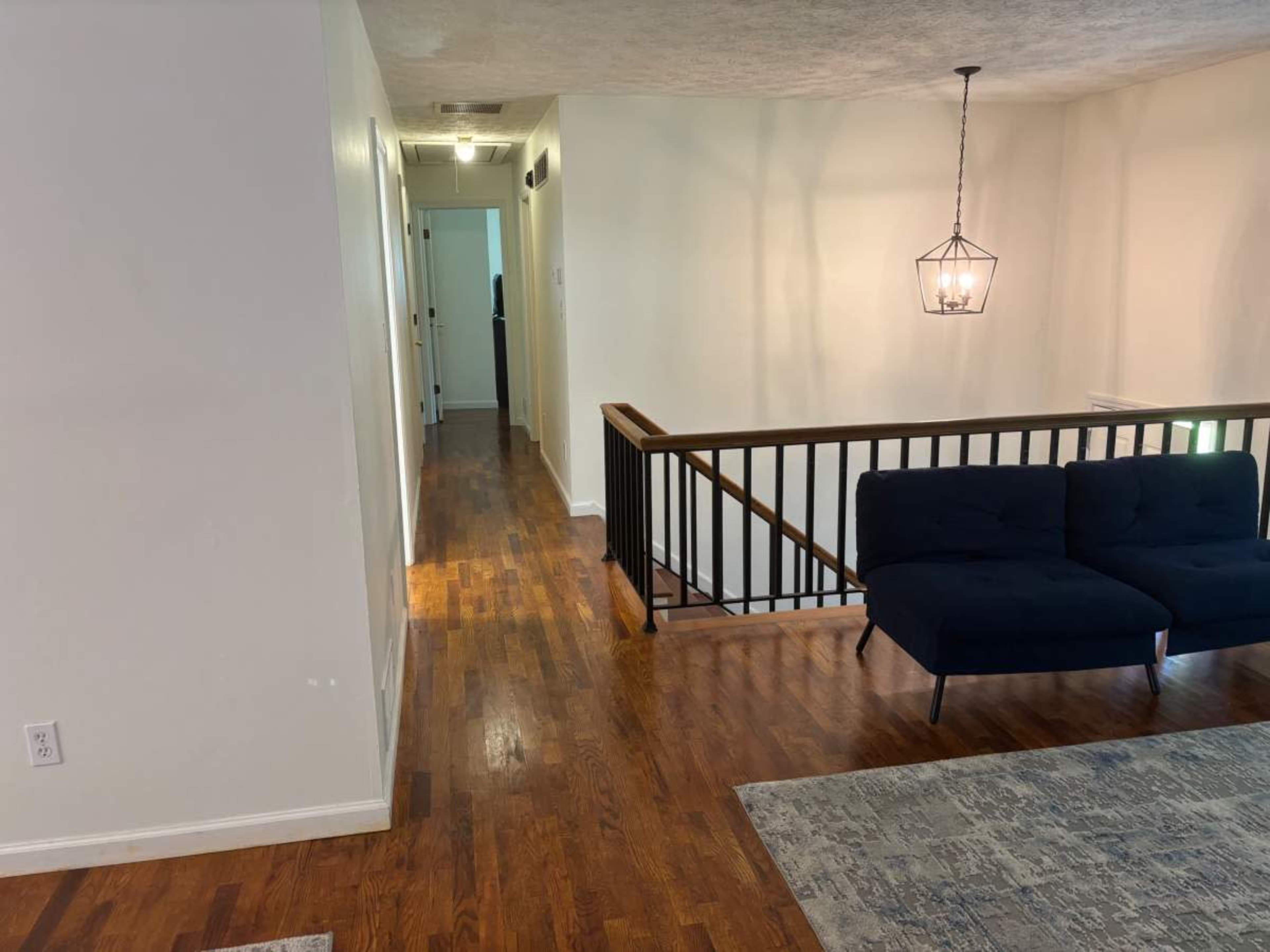 The image shows a living space with hardwood floors, a dark blue couch, and a hallway leading to other rooms, illuminated by a hanging light fixture.