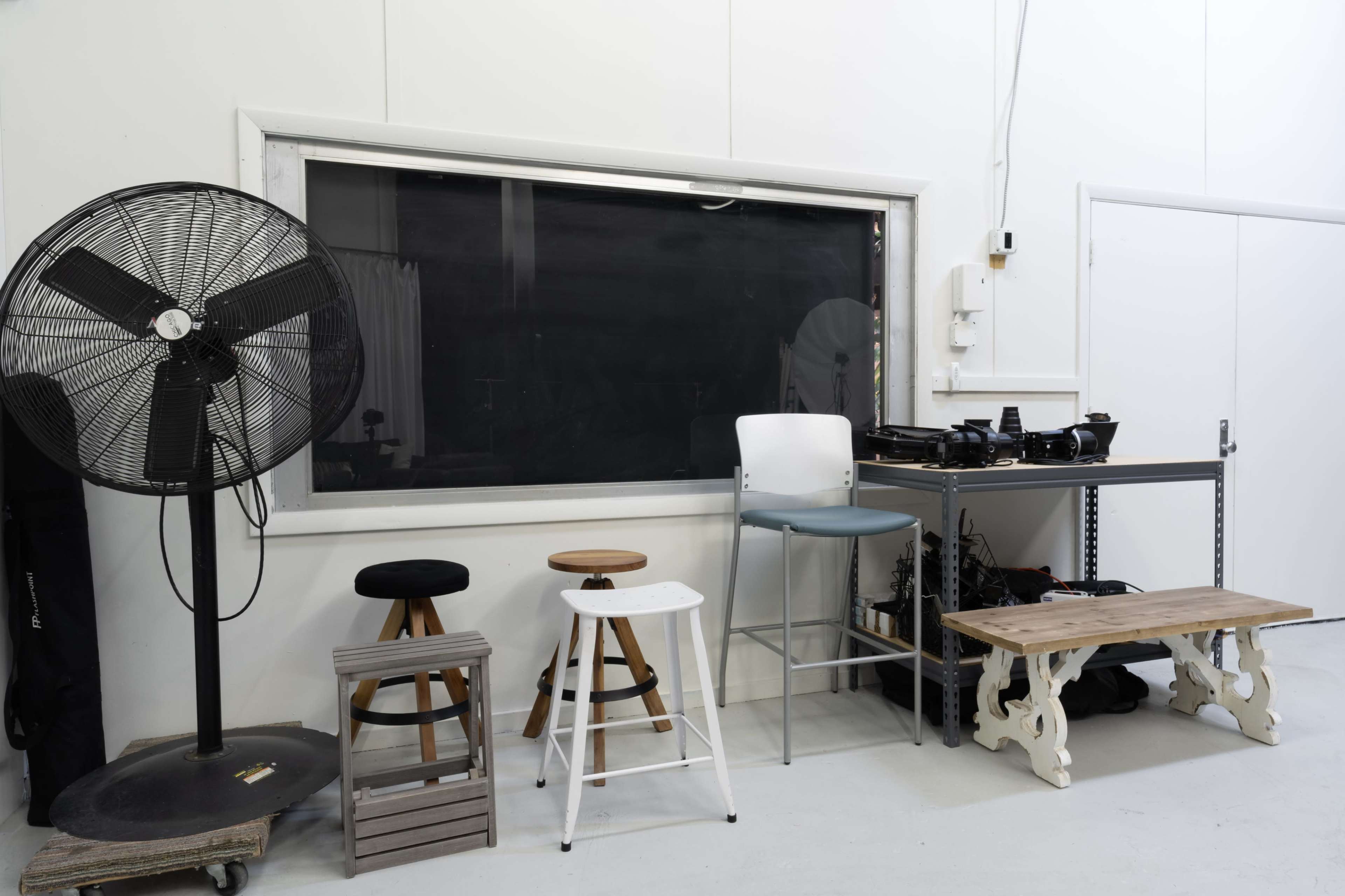 A minimalist room with a large chalkboard, several stools of different heights, a fan, and a wooden bench arranged against white walls.