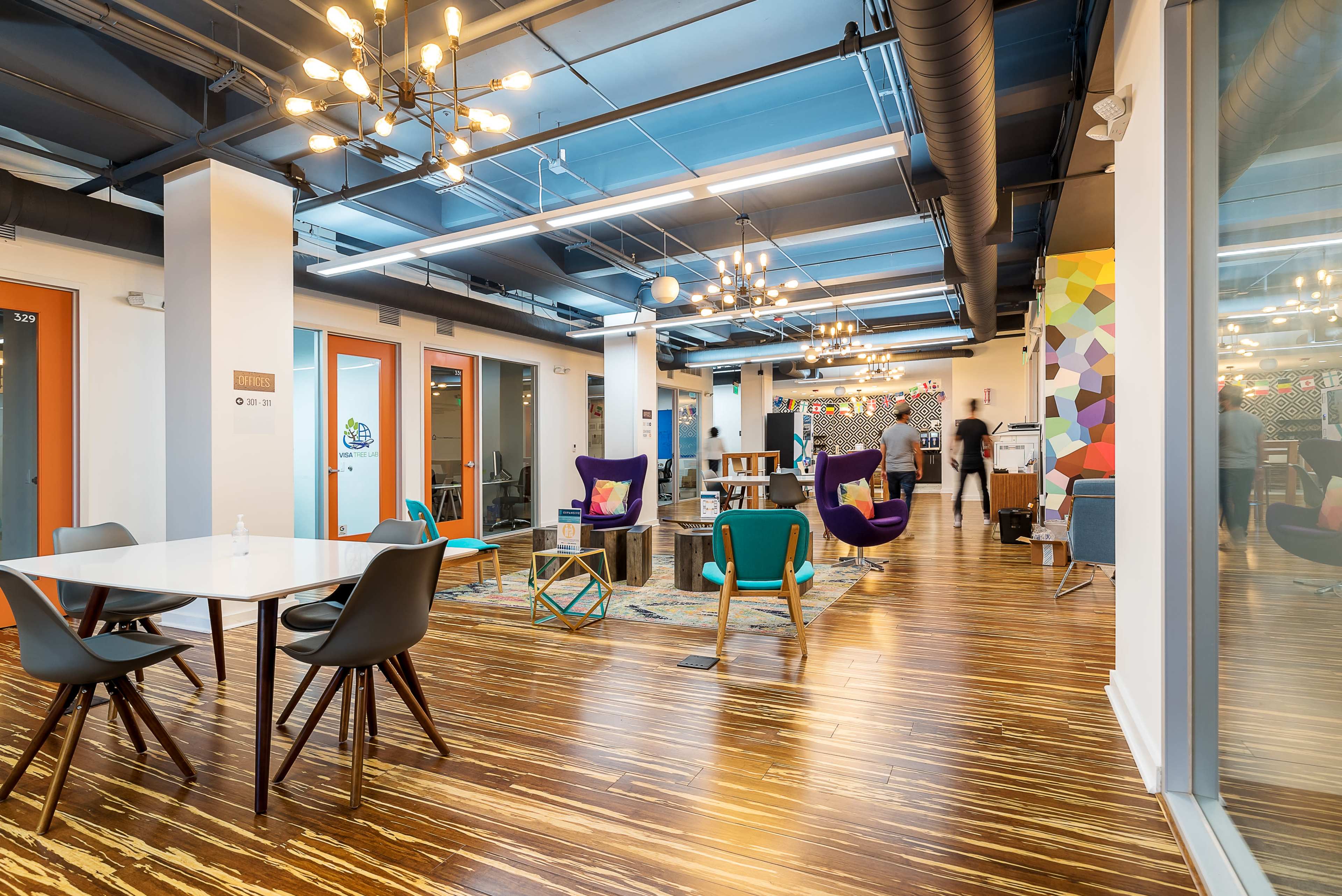 The image shows a modern office space with diverse seating arrangements, wooden flooring, and colorful decor elements.