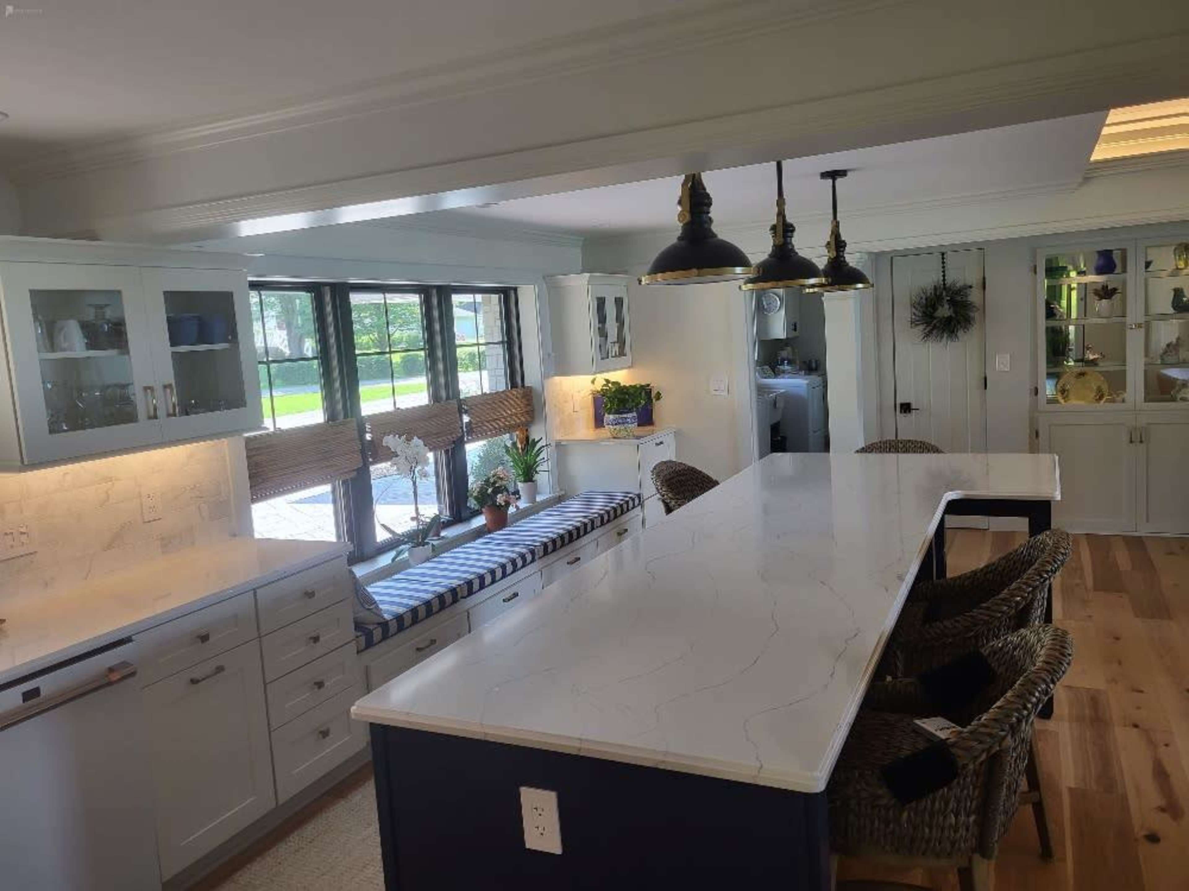 A modern kitchen featuring a large central island with a marble countertop, cabinets with a light color scheme, and a seating area with a cushioned bench near large windows.