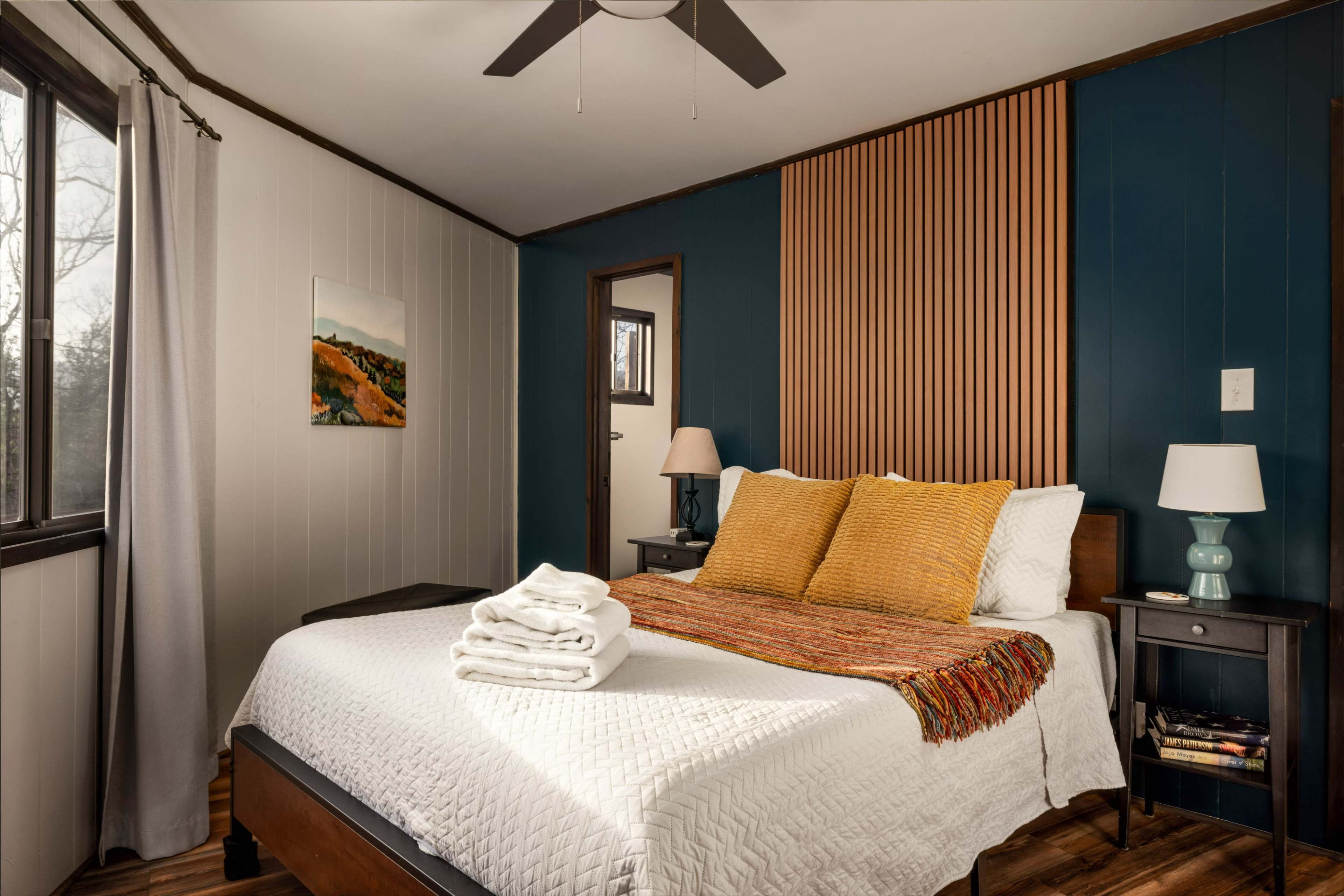The room features a large bed with a white quilt, two yellow pillows, and a decorative throw, set against a blue accent wall with wooden paneling.