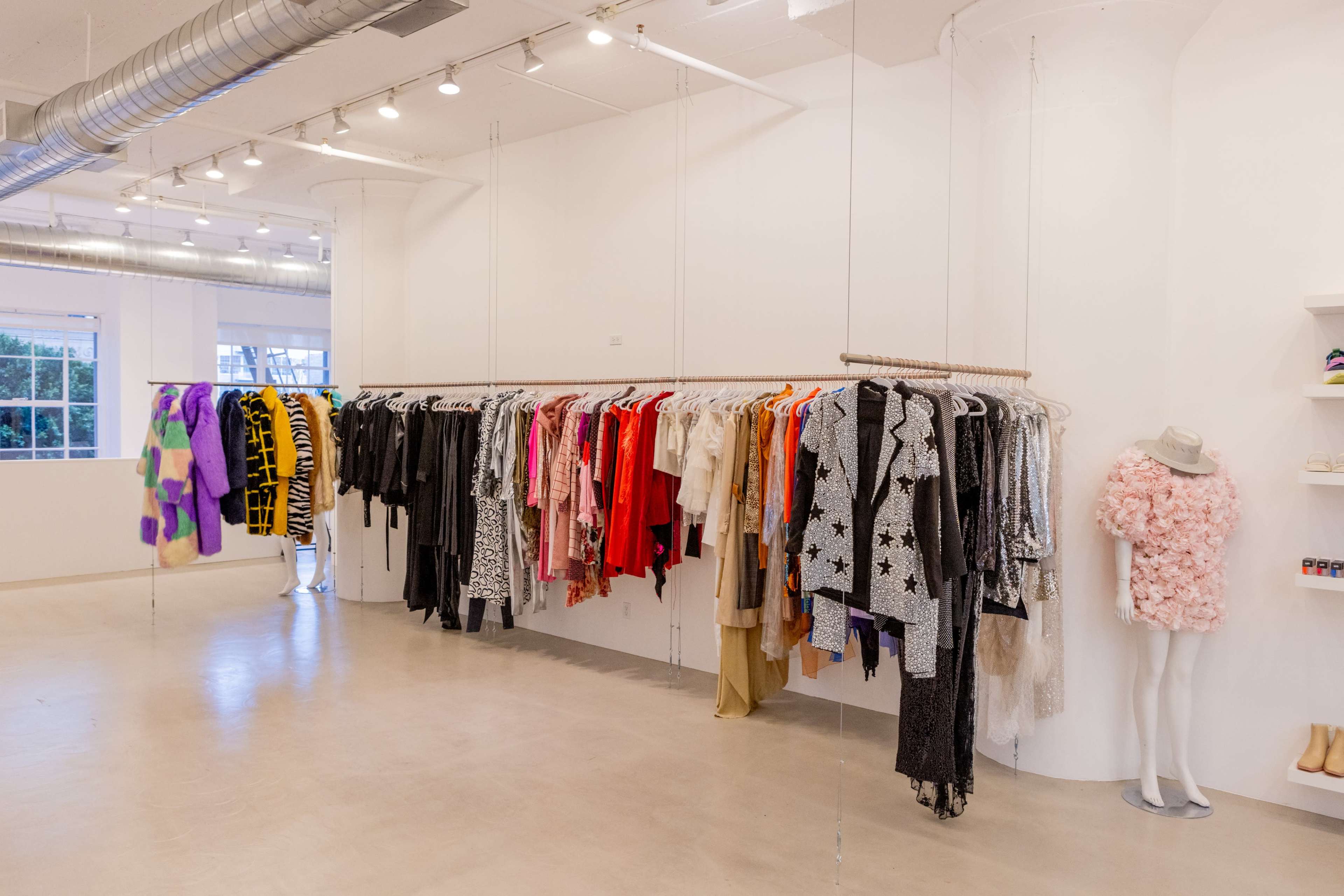 The image shows a clothing store interior with a rack of colorful garments hanging on the left and a mannequin displaying an outfit on the right against white walls.