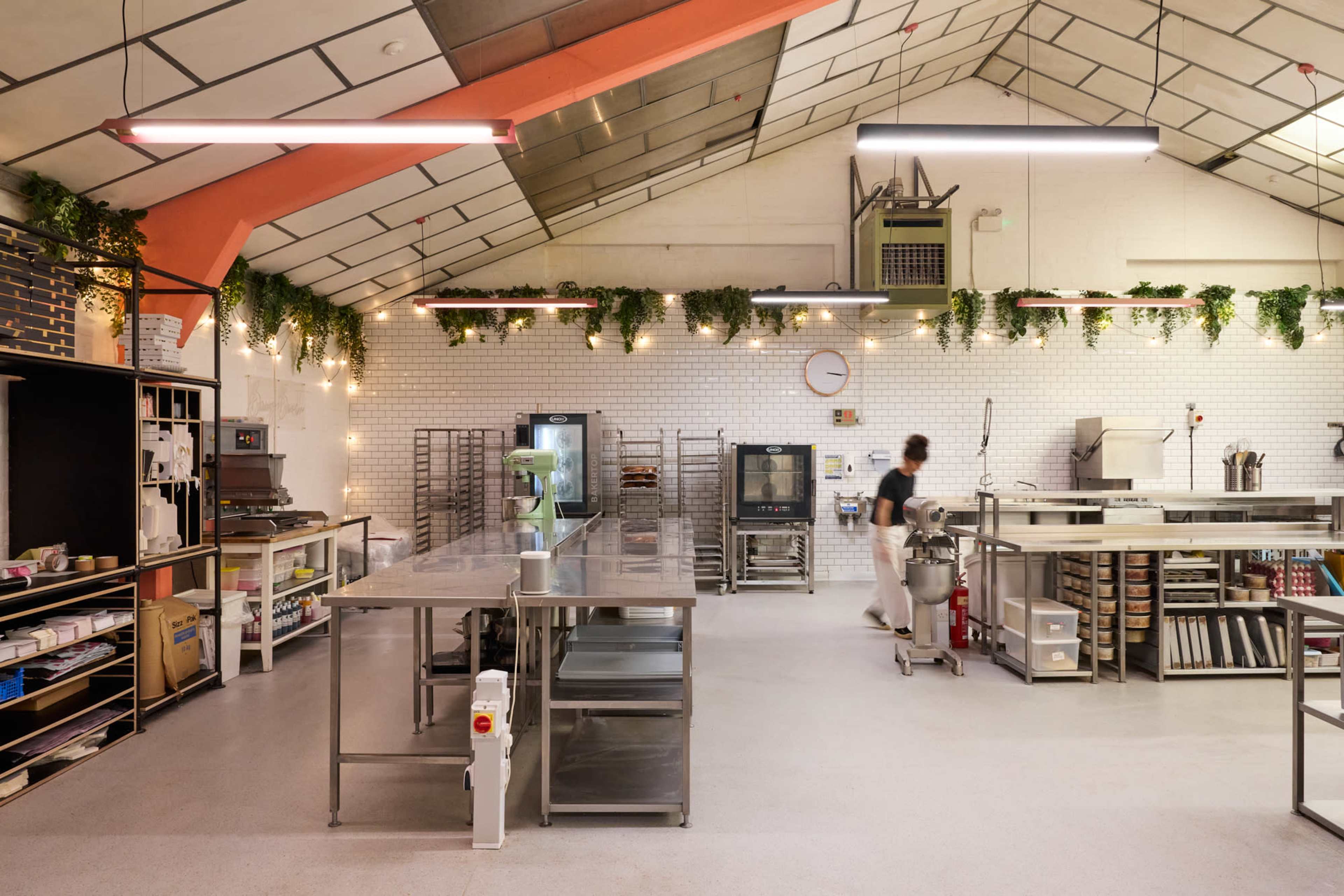 The image shows a spacious commercial kitchen with stainless steel tables, an oven, and overhead greenery, while a person is seen working in the background.